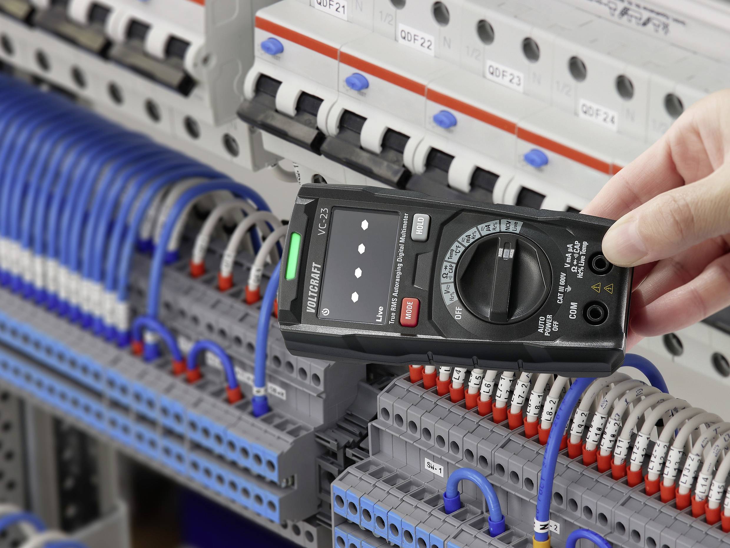 En hand håller ett multimeter framför ett öppet elskåp med blå och grå kablar för att mäta elektriska värden.