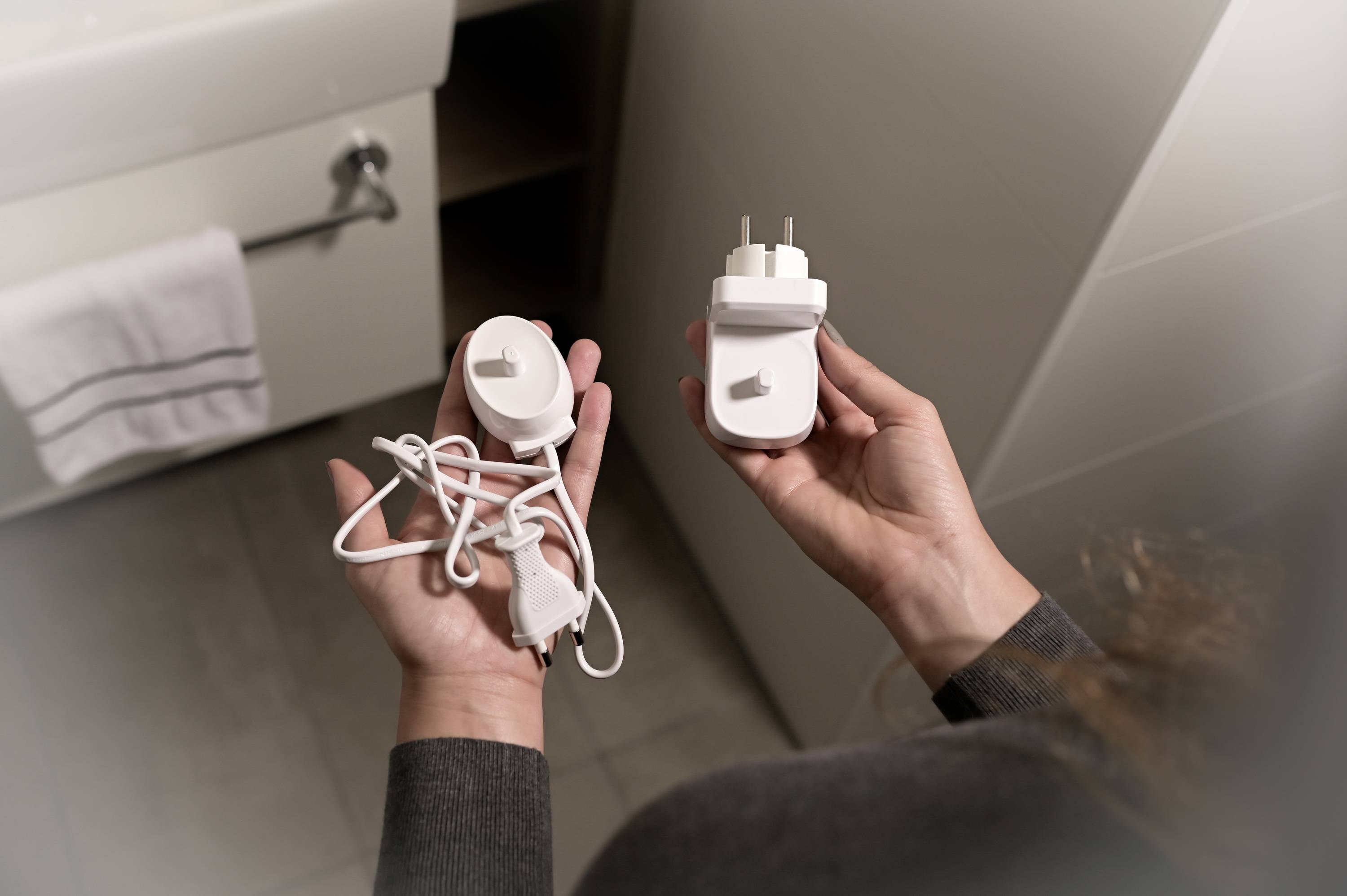 En person håller en vit rakapparat-väggadapter och en lös sladd i händerna, stående i badrummet.