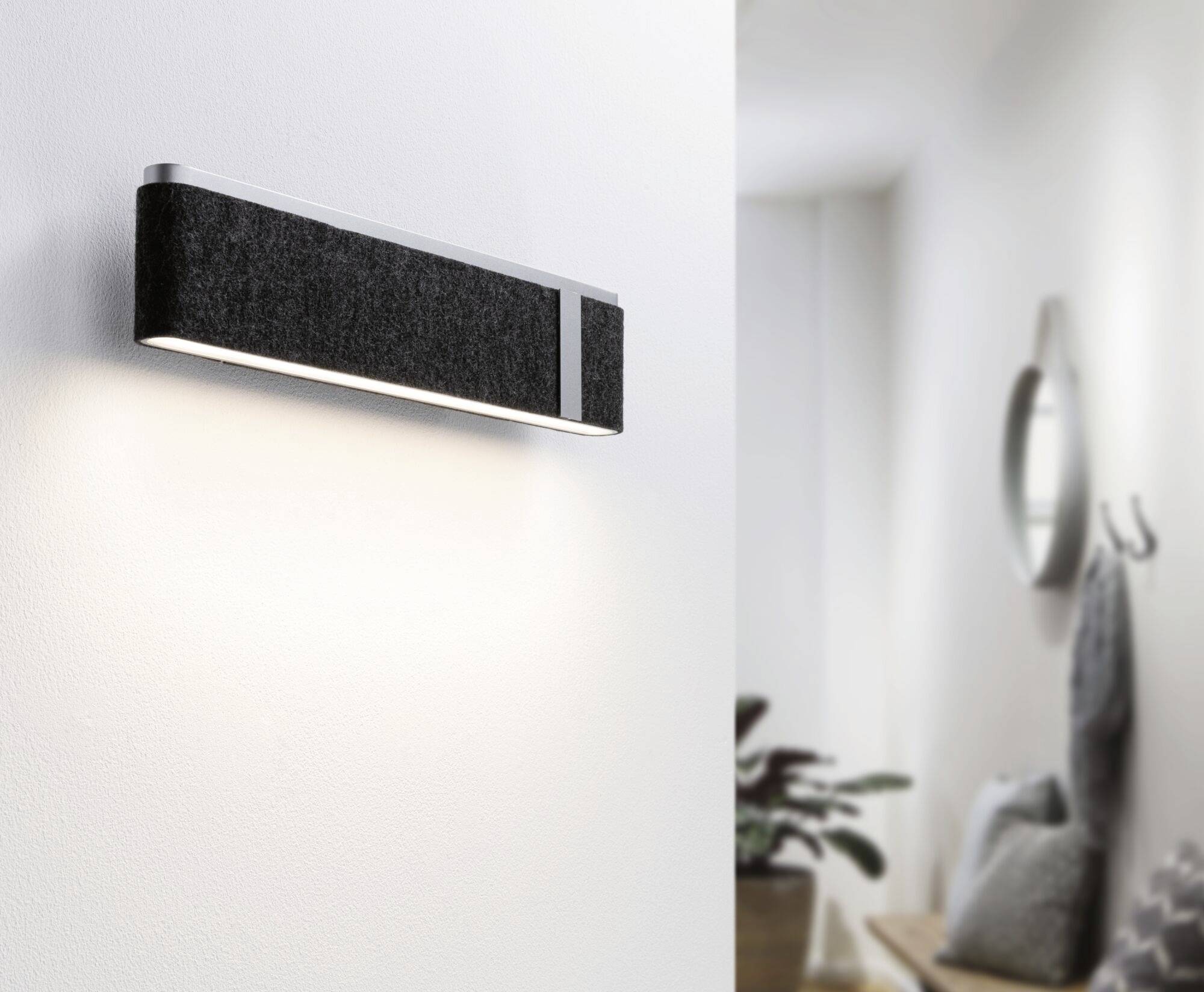 Modern svart vägglampa lyser i ett minimalistiskt inrett rum. I bakgrunden: växt, spegel och klädhängare.