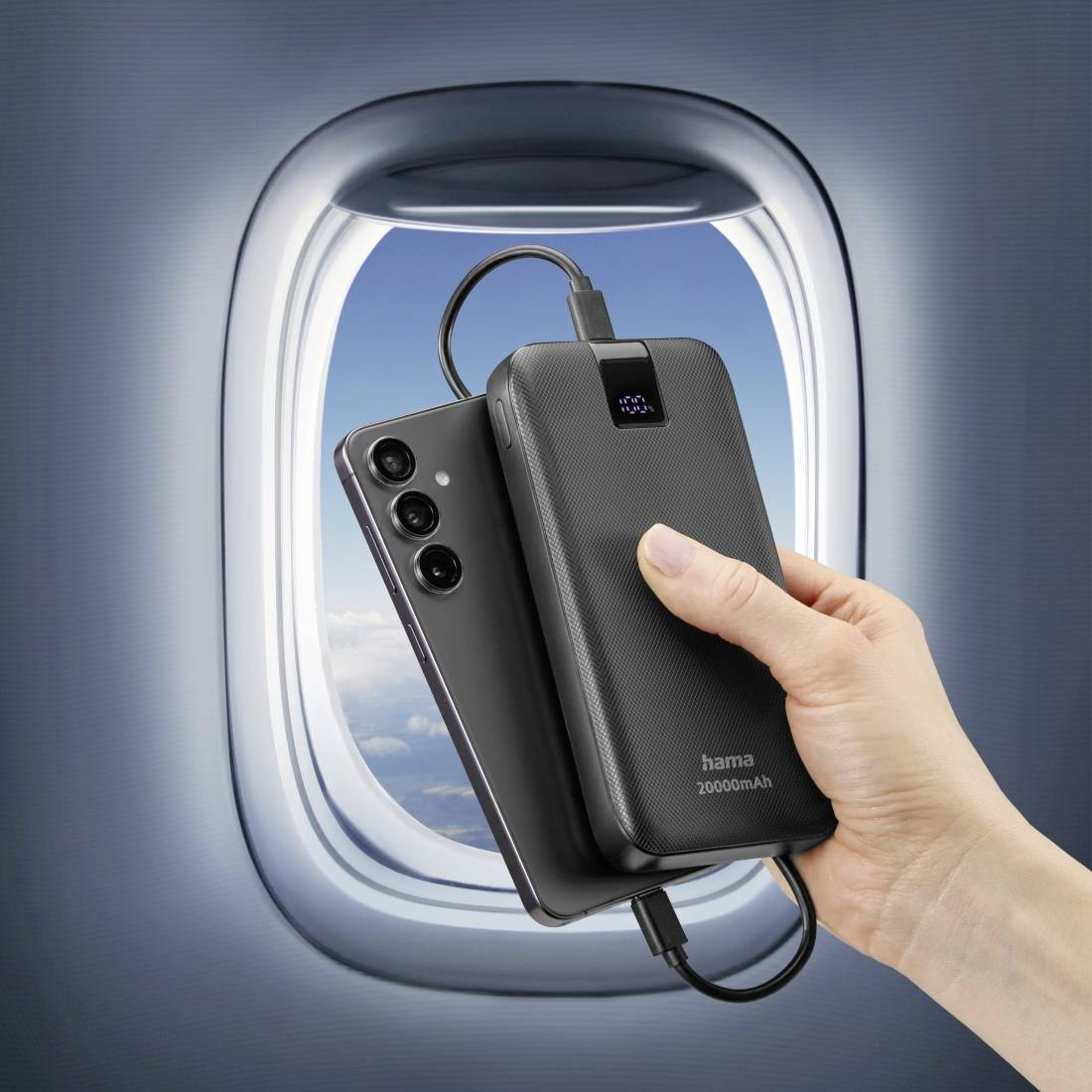 En hand håller en powerbank och en smartphone framför ett flygplansfönster. Båda enheterna är anslutna via en kabel.