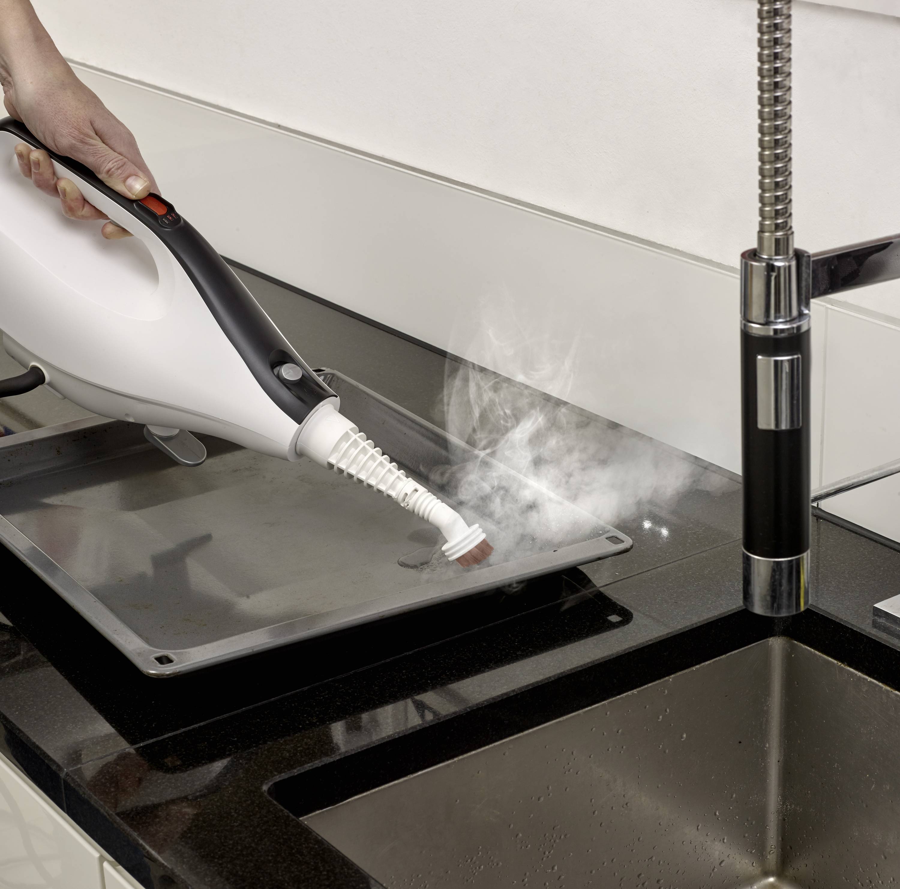 En person rengör en spishäll med en ångstädare. Ångan avlägsnar smuts från ytan bredvid en rostfri diskho.