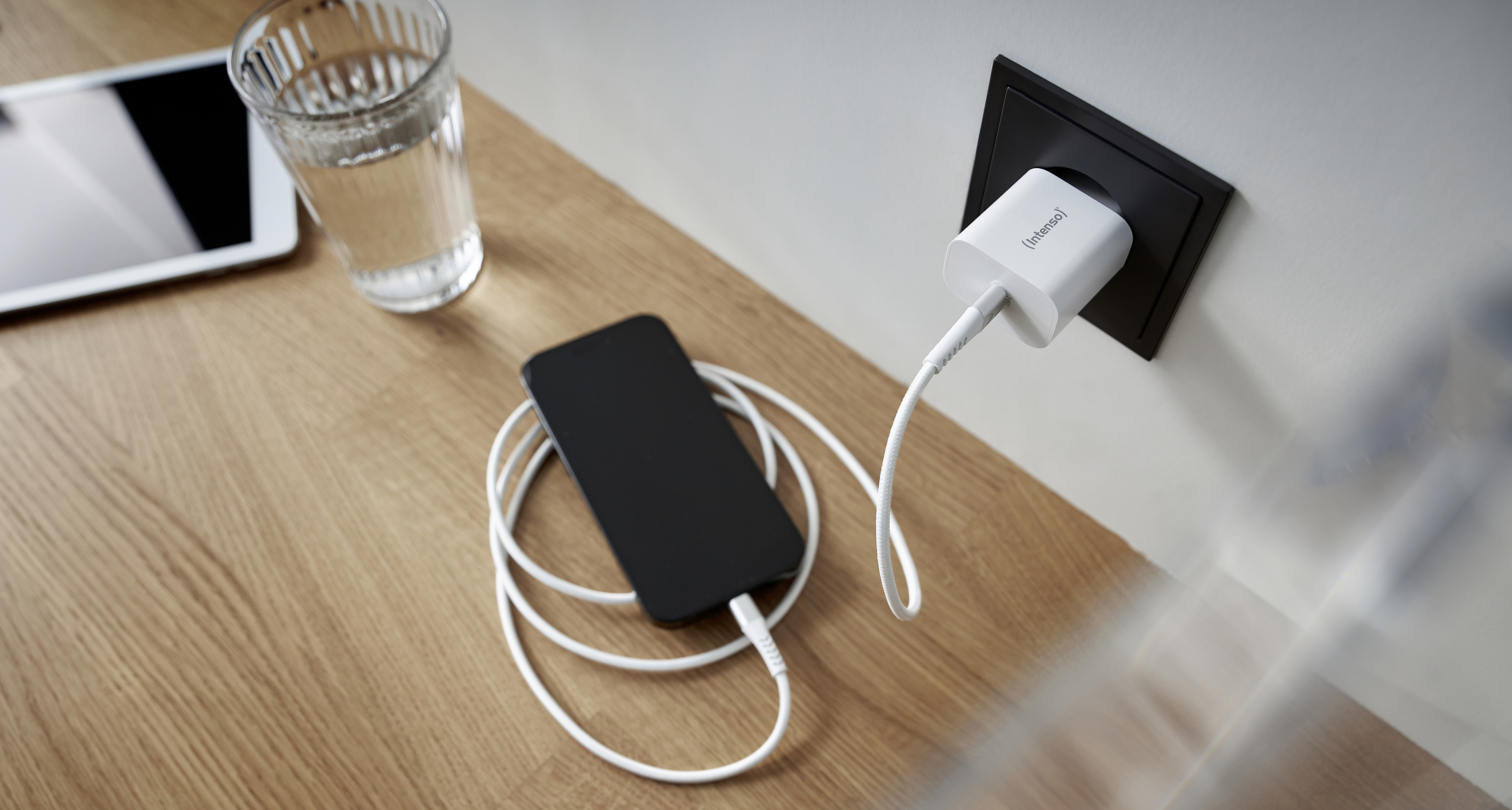 En smartphone ligger på ett träbord och laddas via en vit kabel i ett eluttag. Bredvid finns ett glas vatten och en surfplatta.