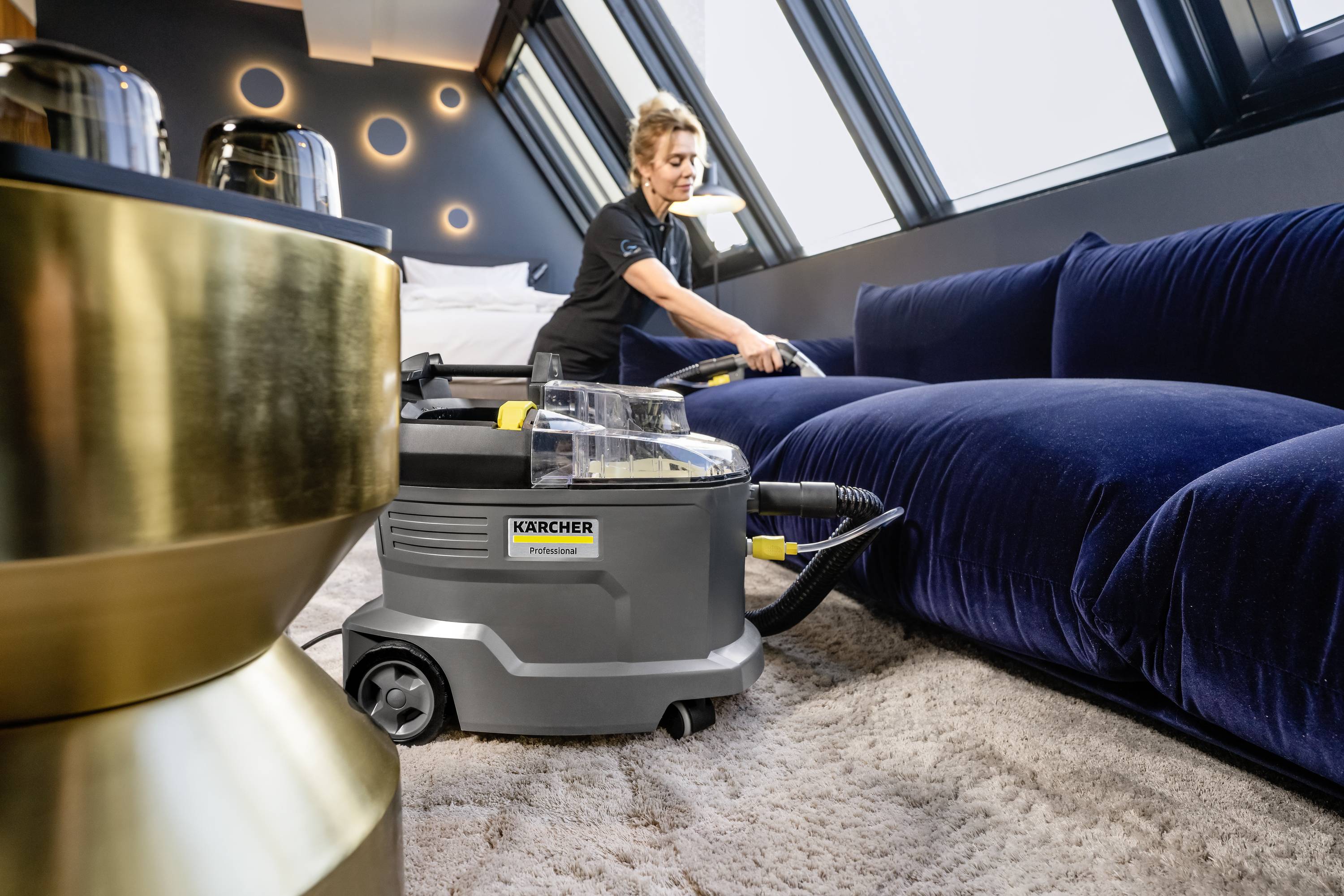 En person rengör en blå sammetssoffa med en professionell mattborttagare i ett modernt, välbelyst rum med snedställda fönster.