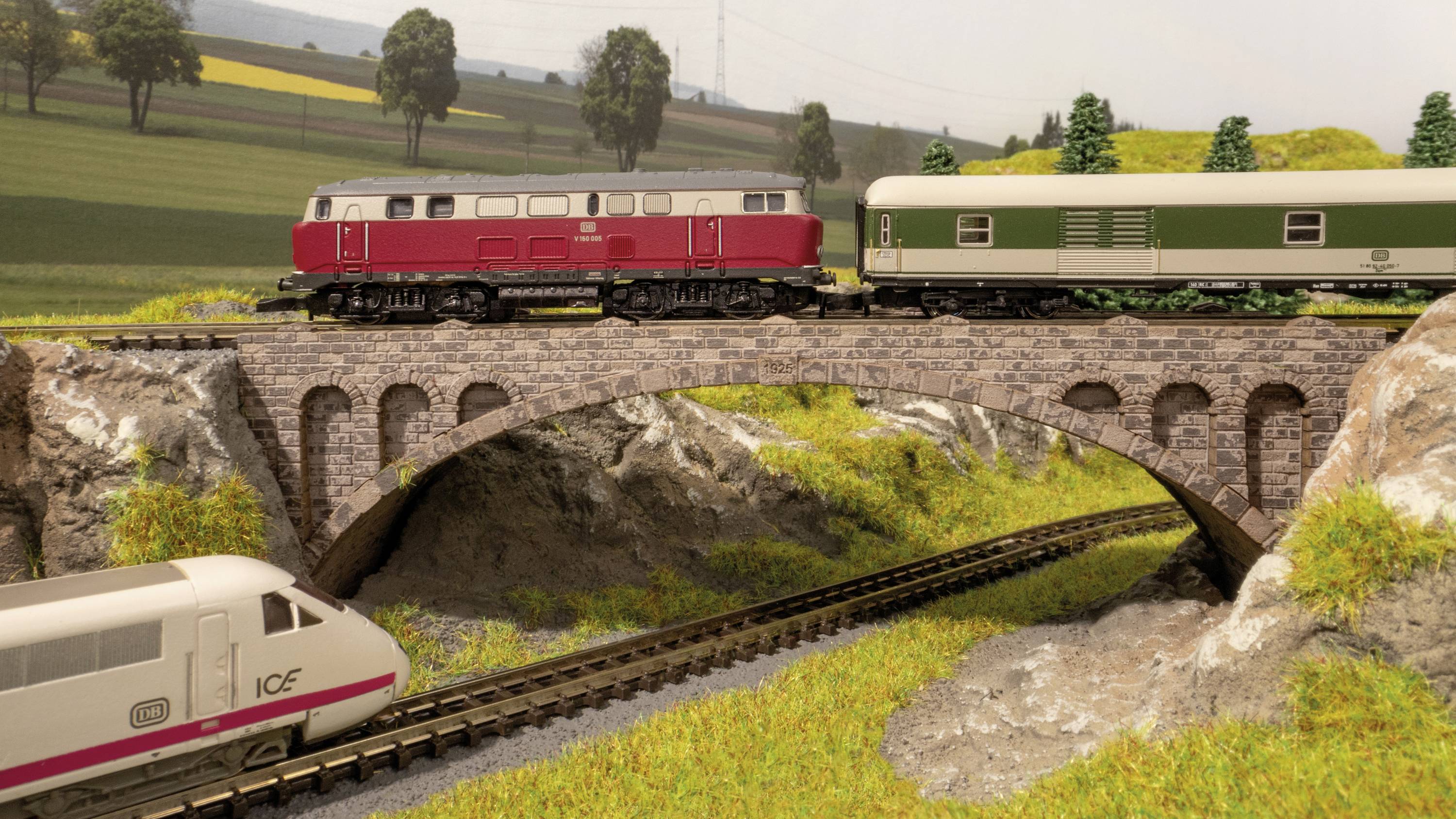 En modelljärnvägslandskap visar ett rött och ett grönt lok på en bro. Under denna kör ett ICE-tåg på ett annat spår.