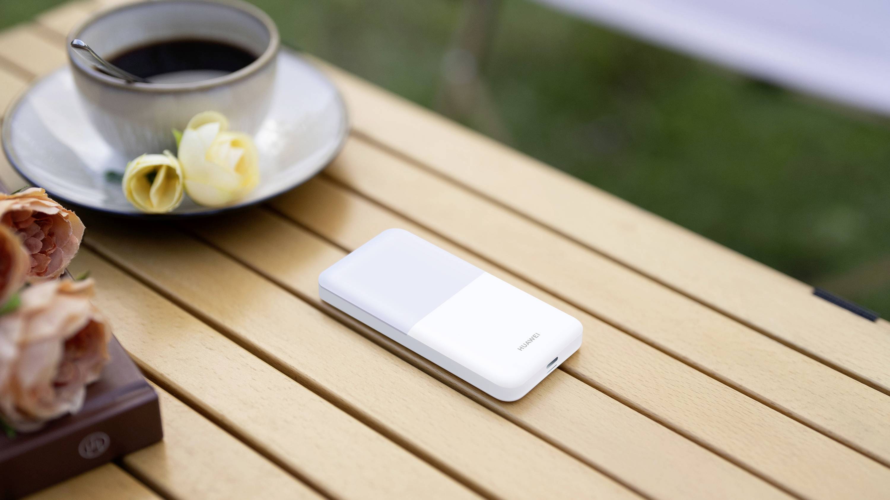 En mobiltelefon ligger på ett träbord bredvid en kopp kaffe och gula rosor. I bakgrunden syns en grön omgivning.
