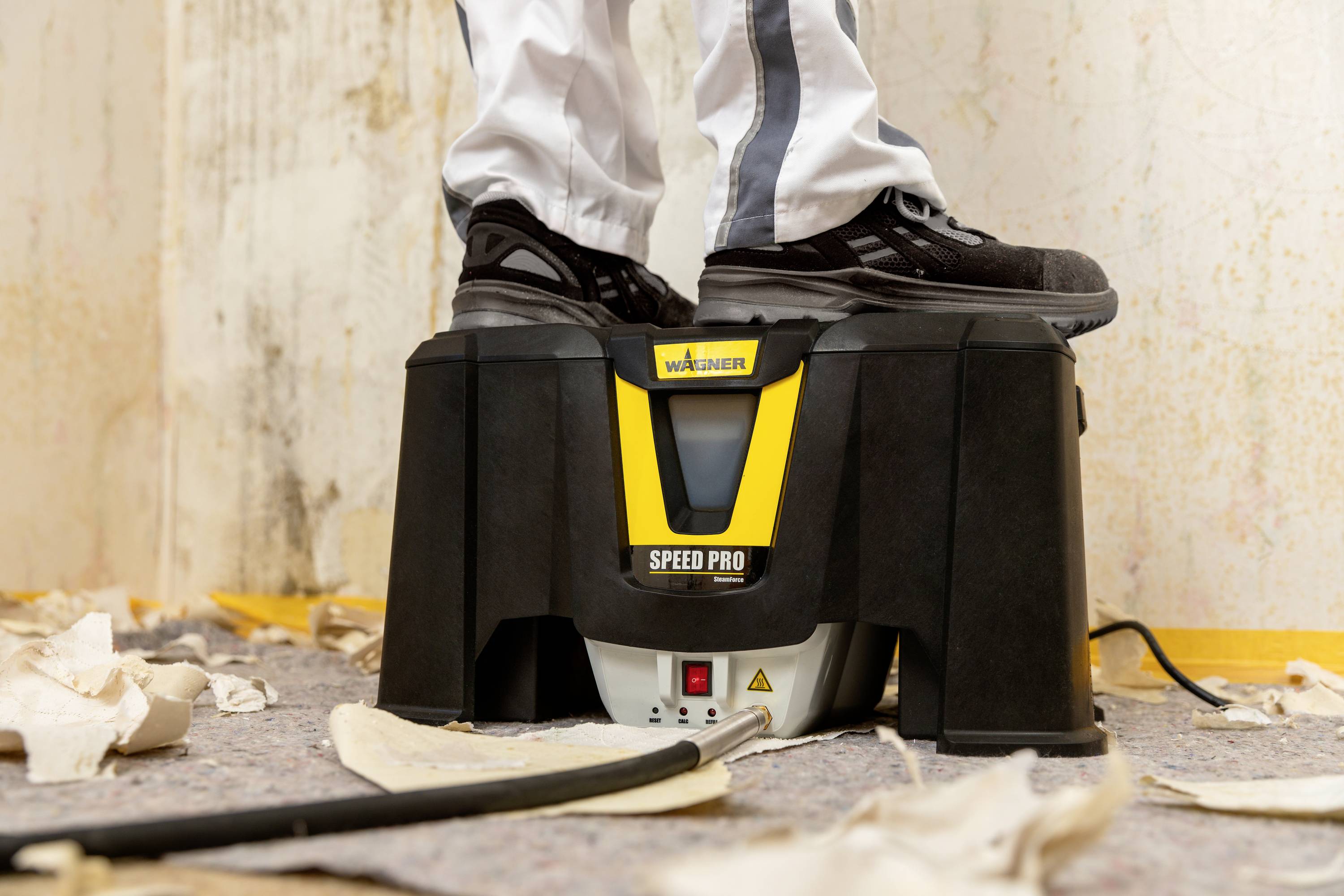 En person står på en svart och gul apparat märkt 'Speed Pro', omgiven av trasiga tapetbitar.