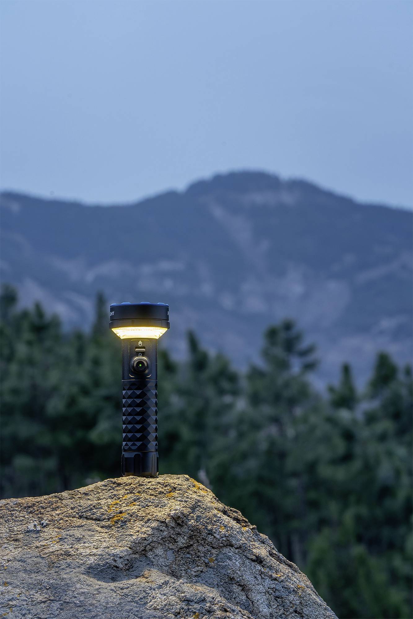 Ficklampa lyser på en klippa i skymningen, med skogbeklädda berg i bakgrunden.