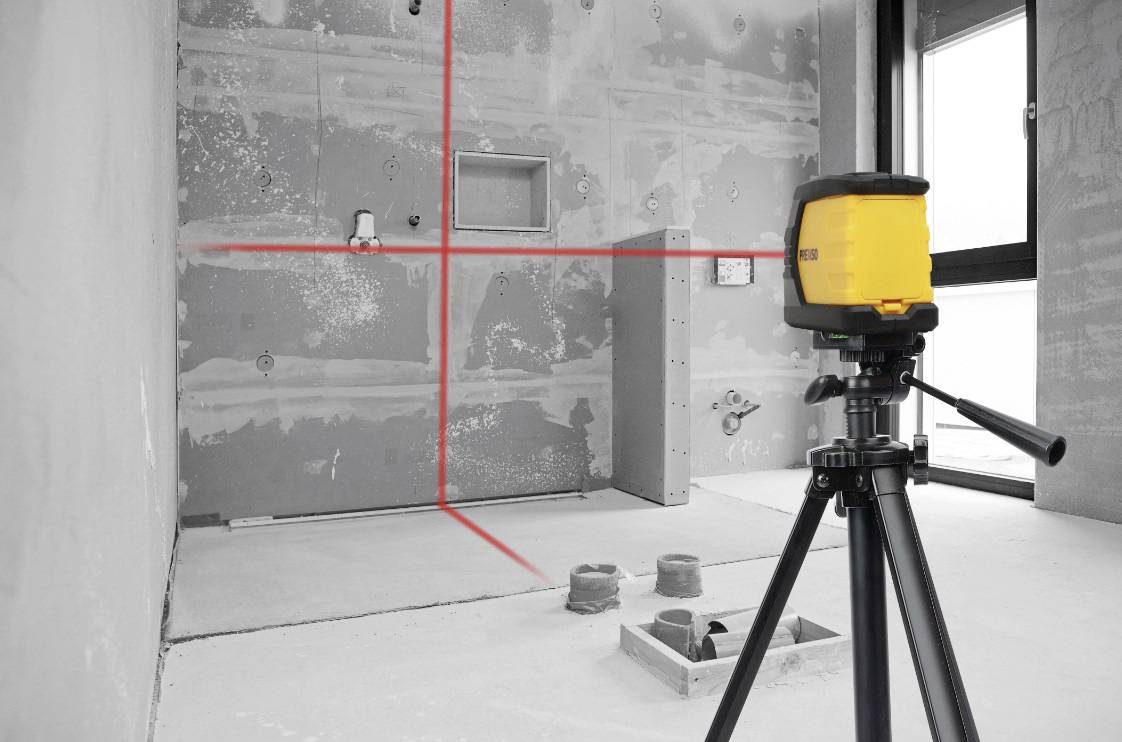En lasernivåinstrument på ett stativ projekterar röda linjer på en grå vägg på en byggarbetsplats; den visar exakta mätlinjer.