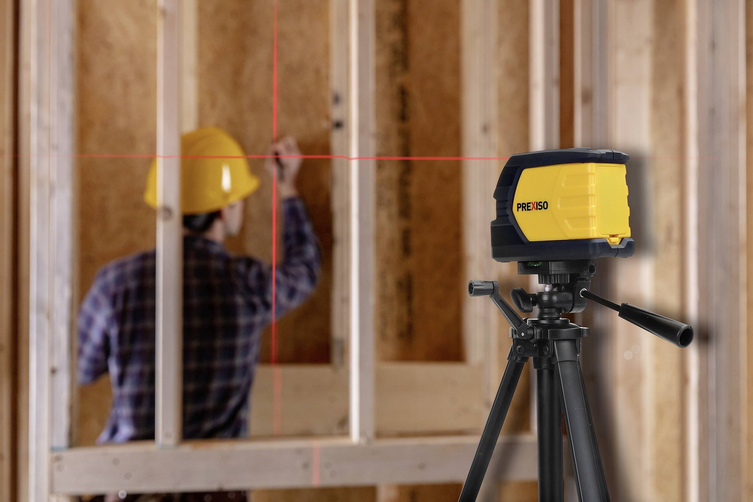 Byggnadsarbetare med skyddshjälm justerar träram i ett byggprojekt. I förgrunden ett lasernivåinstrument monterat på ett stativ.