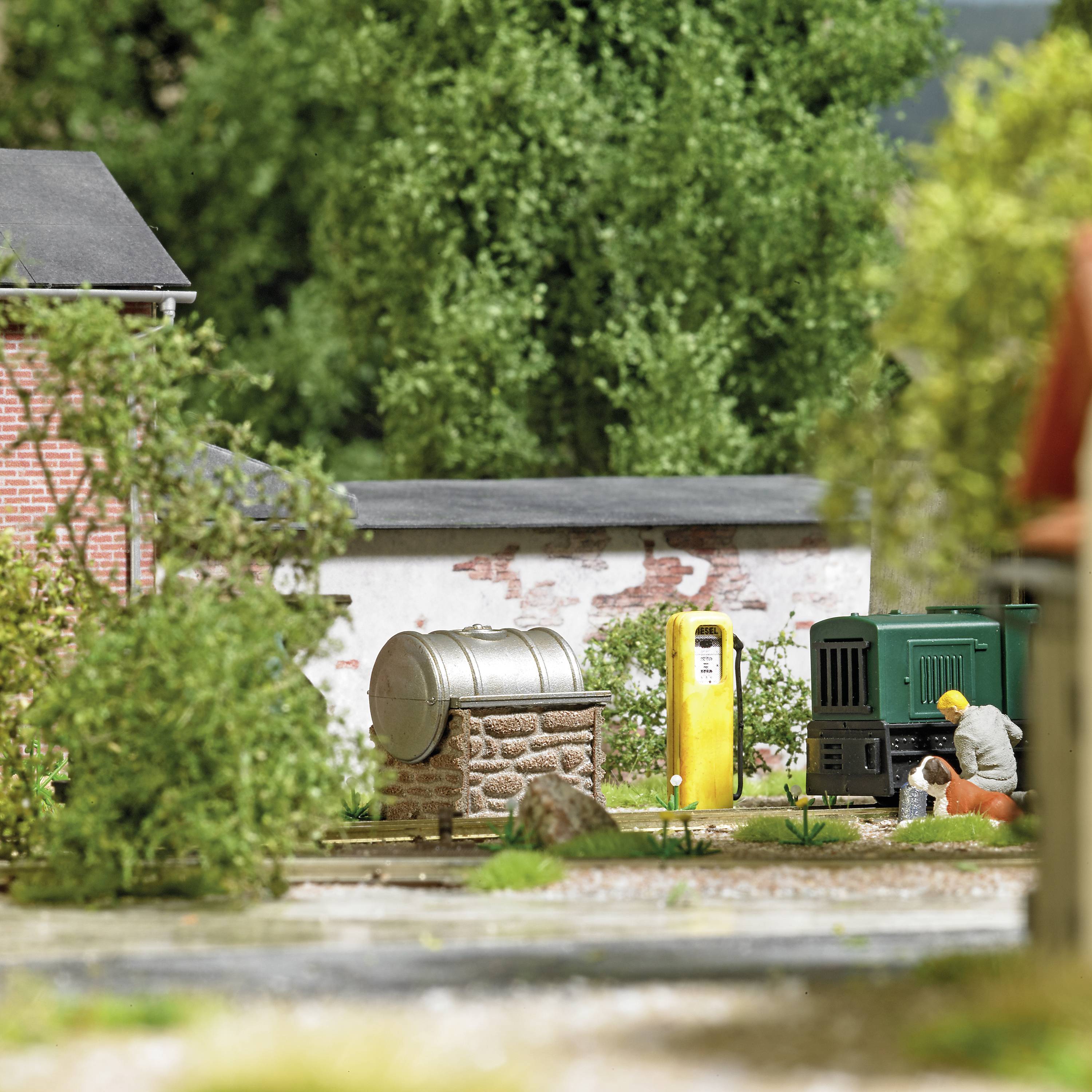 En miniatyrscen med en gul bensinstationsmodell och en modelbyggnadsarbetare som reparerar något på ett grönt maskinartat objekt.