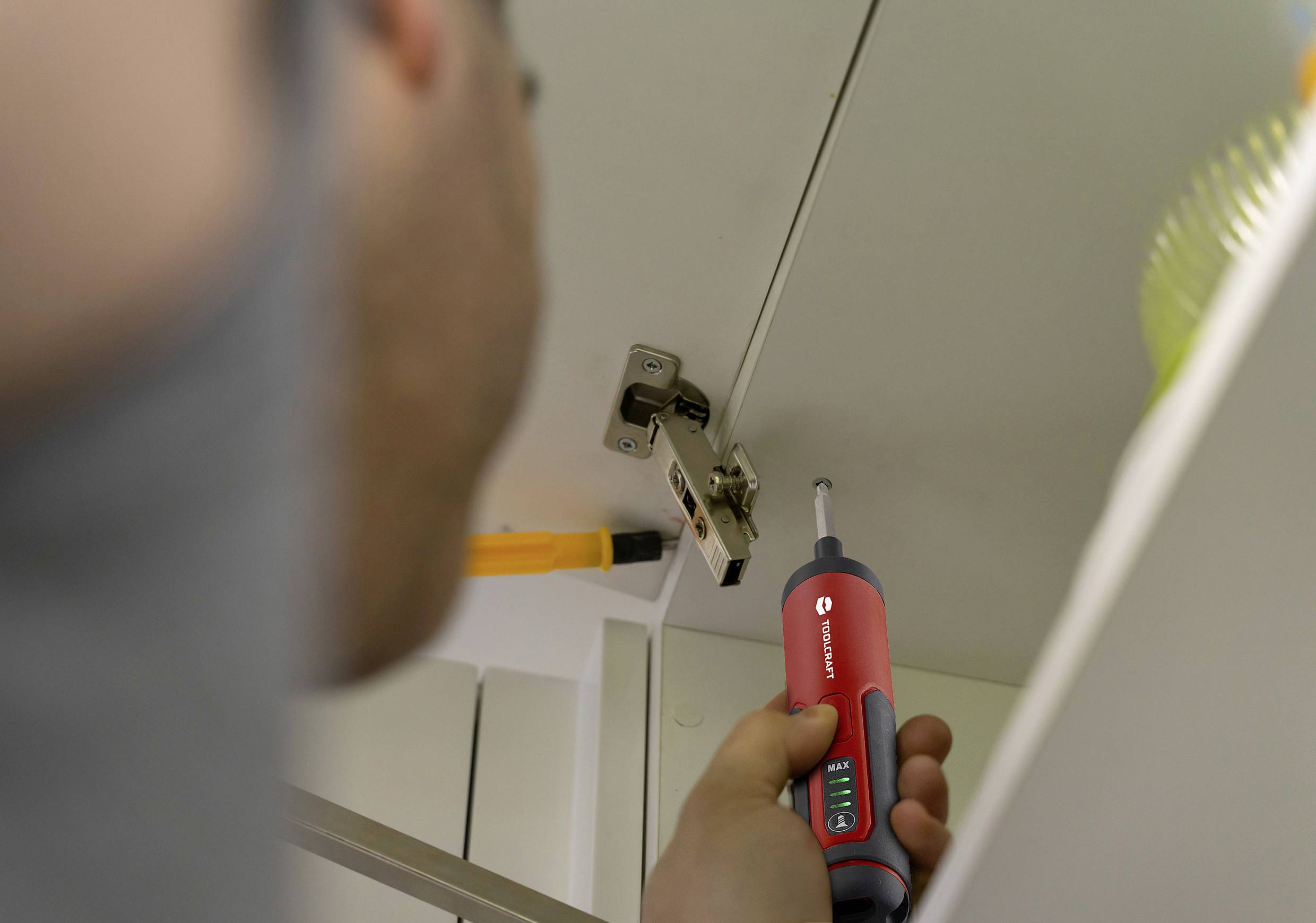 En person reparerar ett gångjärn på ett skåp med en röd skruvmejsel. Skåpet står öppet.
