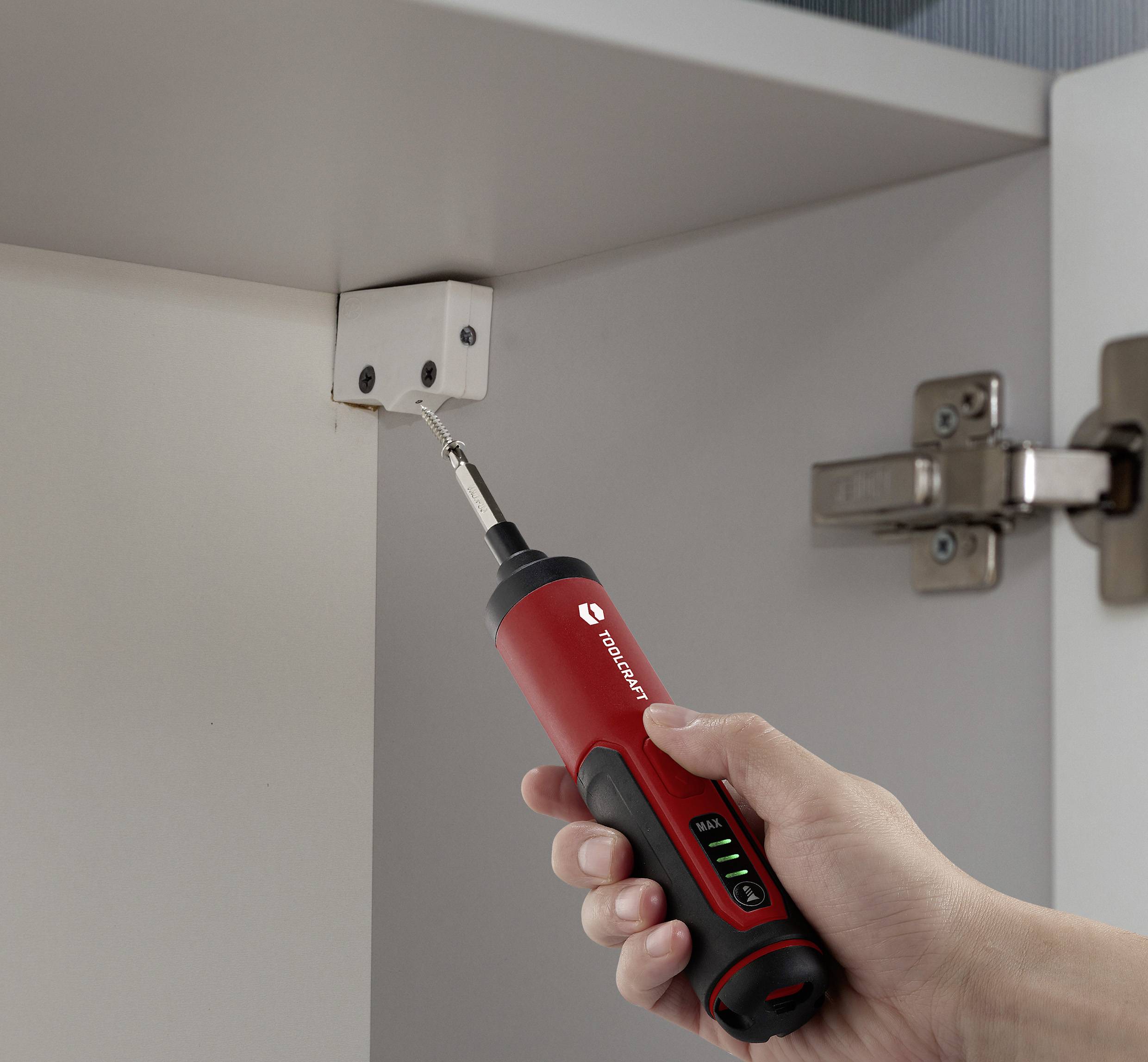 Hand håller en sladdlös skruvdragare och drar åt en skruv i gångjärnet på en vit skåpdörr. Verktyget visar varumärkeslogotyp och batterinivå.