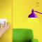 En hand vrider en vit dimmer på en gul vägg. Bredvid finns en violett lampa och en grön stol.