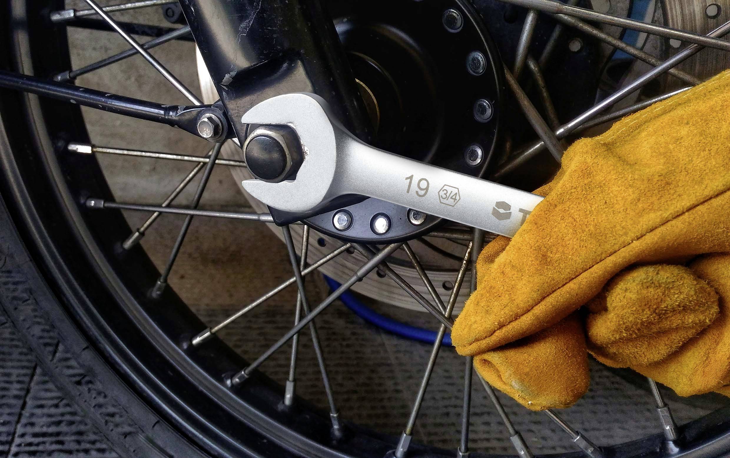 En hand iklädd en gul handske håller en skiftnyckel som är fäst vid en skruv på ett cykel- eller motorcykelhjul.