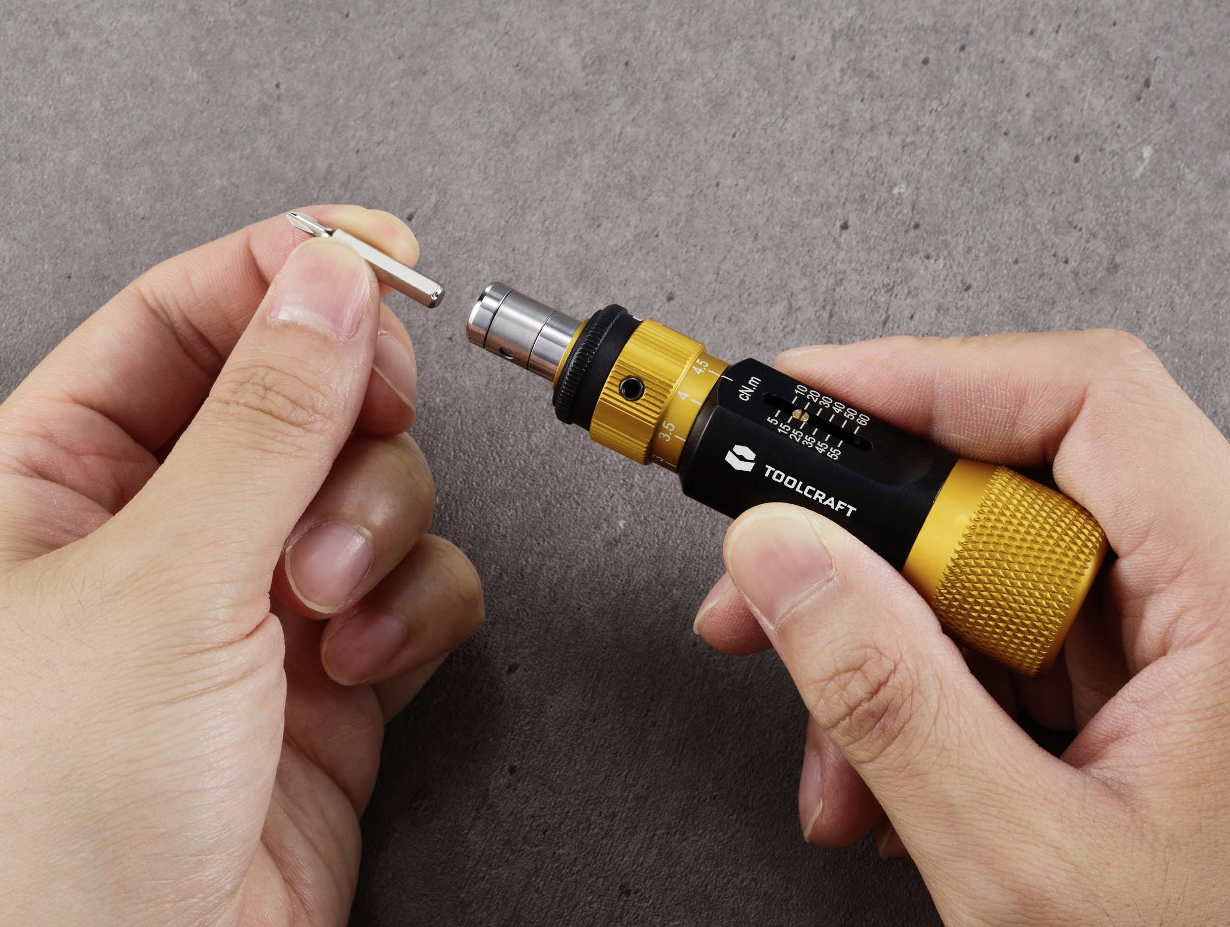 En person håller en gul skruvmejsel och en liten skruv mot en grå bakgrund.