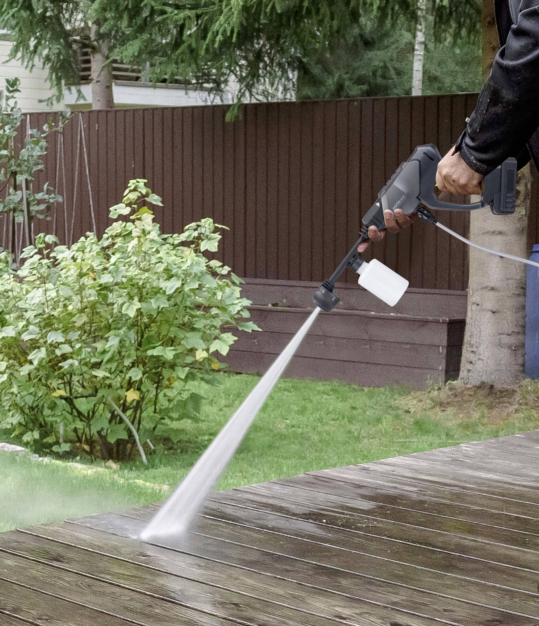 En person rengör en träterrass med en högtryckstvätt; i bakgrunden syns buskar och ett staket.