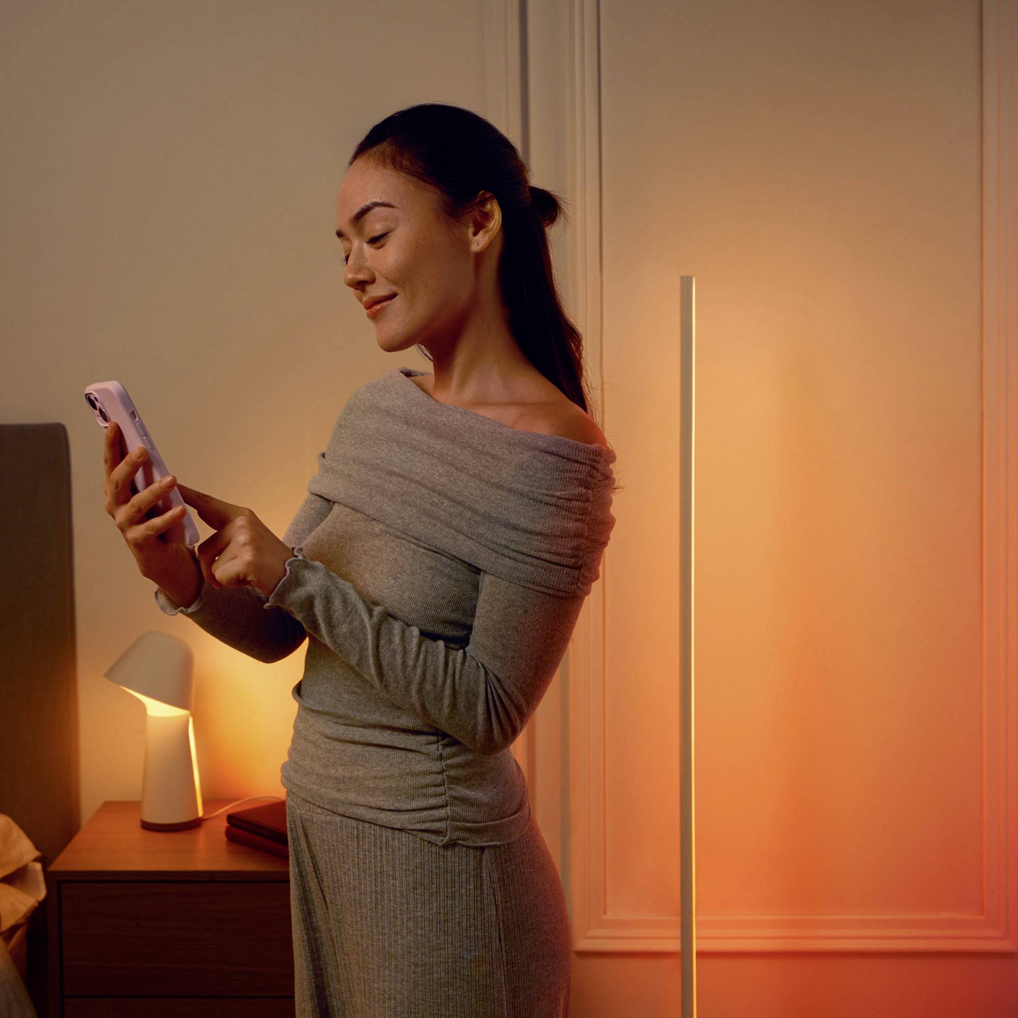 En kvinna i bekväm klädsel står i ett mjukt upplyst sovrum och ler medan hon tittar på sin mobiltelefon.