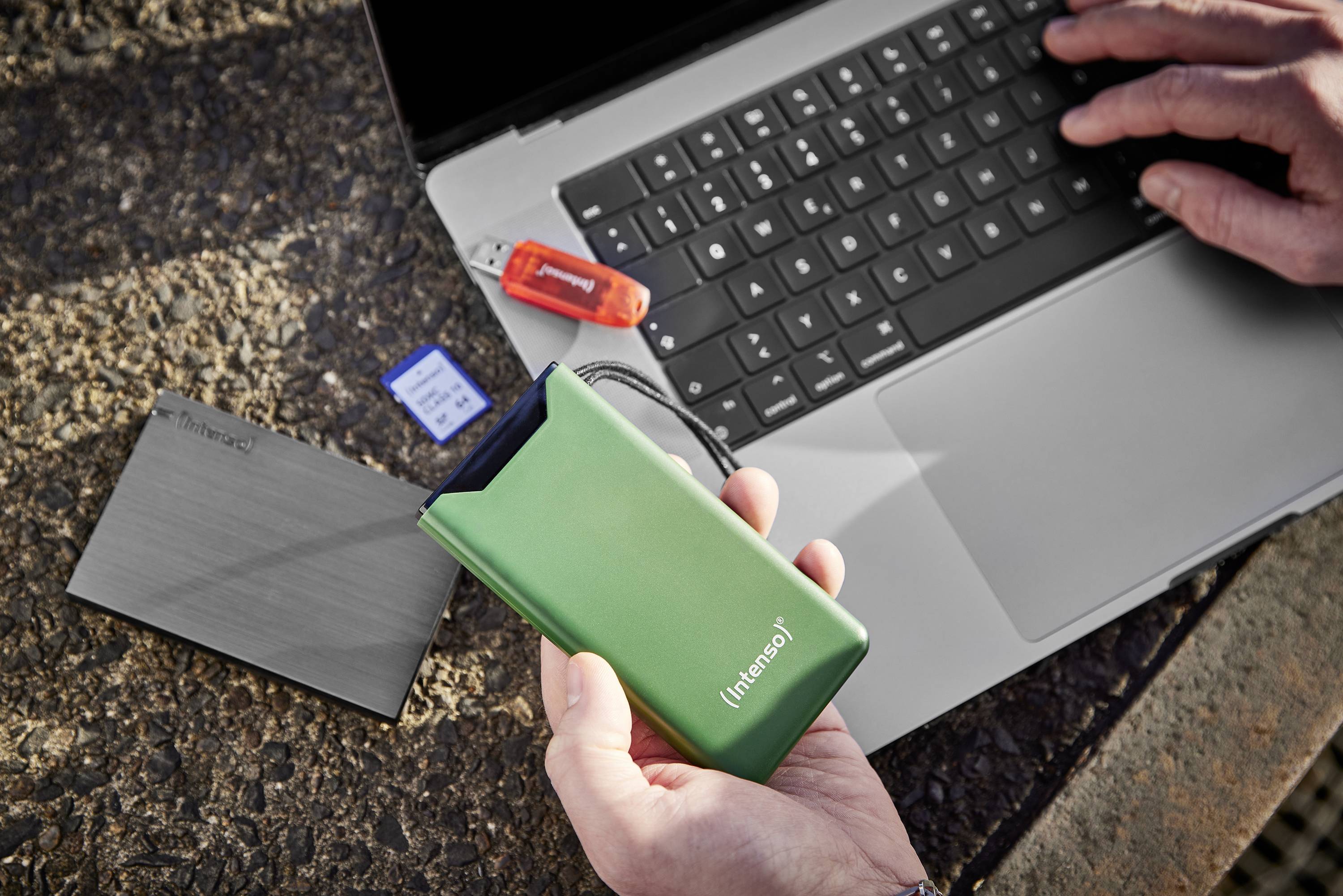 En person håller en grön extern hårddisk framför en bärbar dator. Bredvid ligger ett USB-minne och ett blått SD-kort på ett bord.