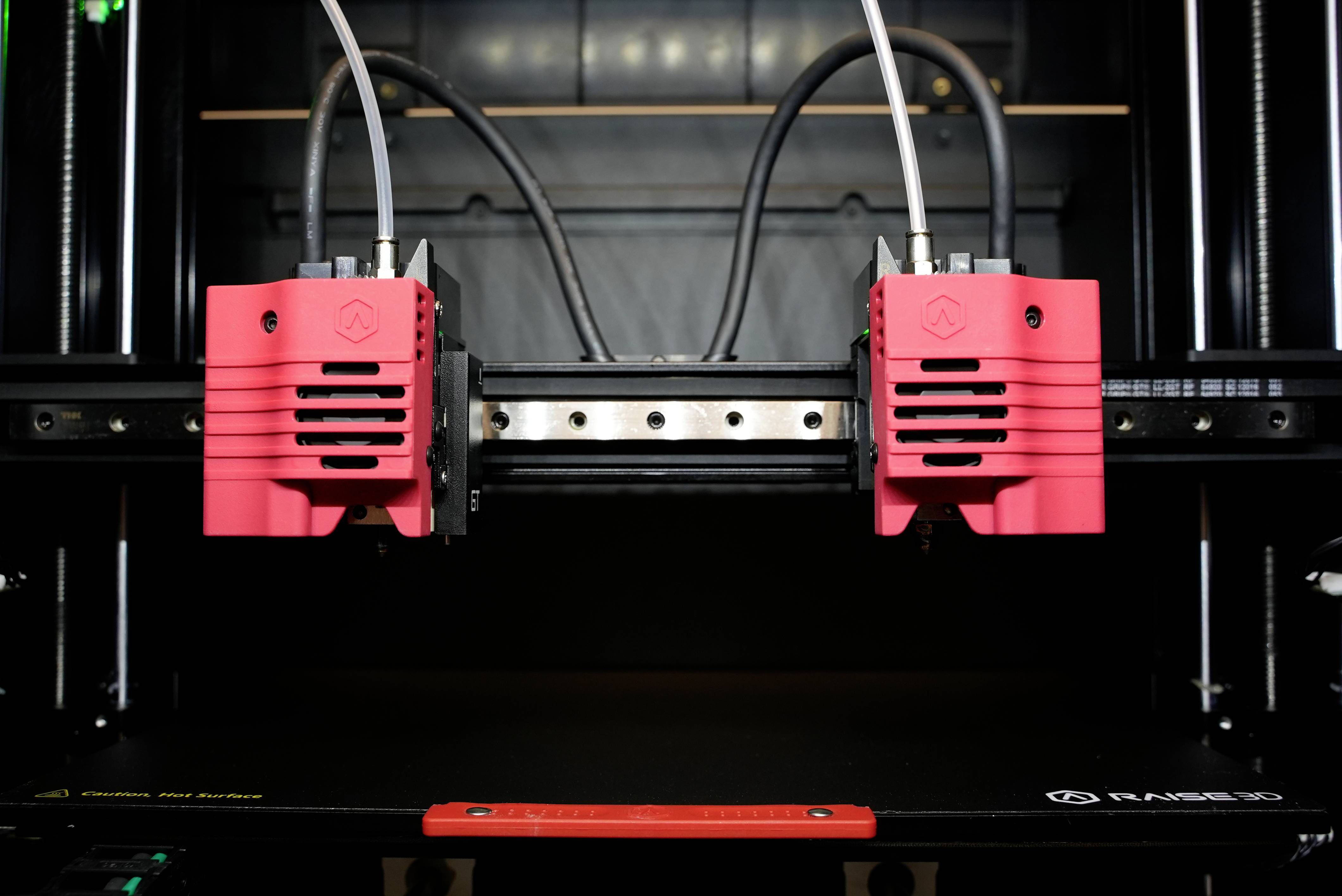 En 3D-skrivare med två röd-svarta extruderare skriver ut på en svart plattform. I bakgrunden syns kablar.