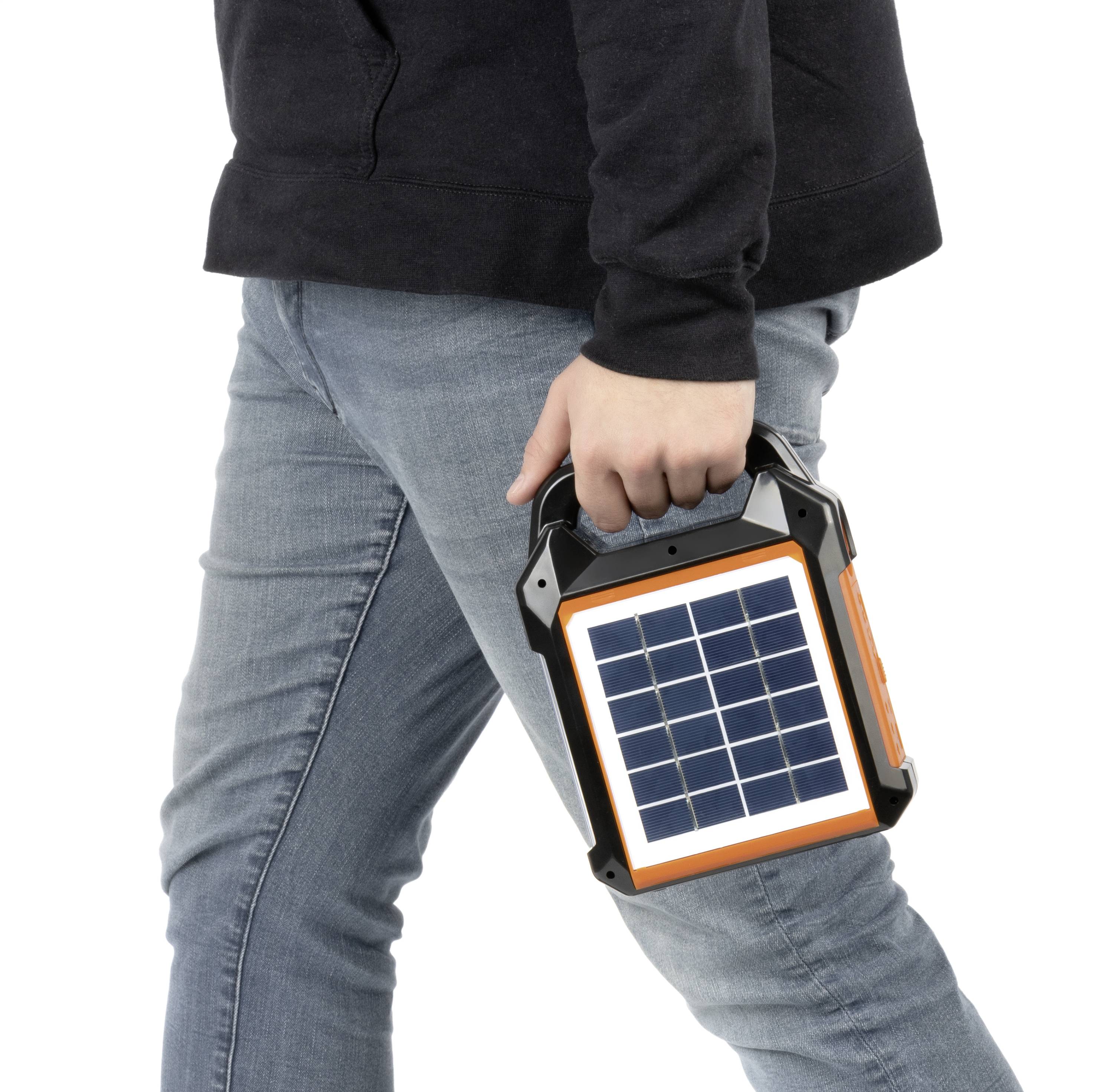 En person i jeans bär en bärbar solargenerator i handen. Enheten har ett solpanel på framsidan.