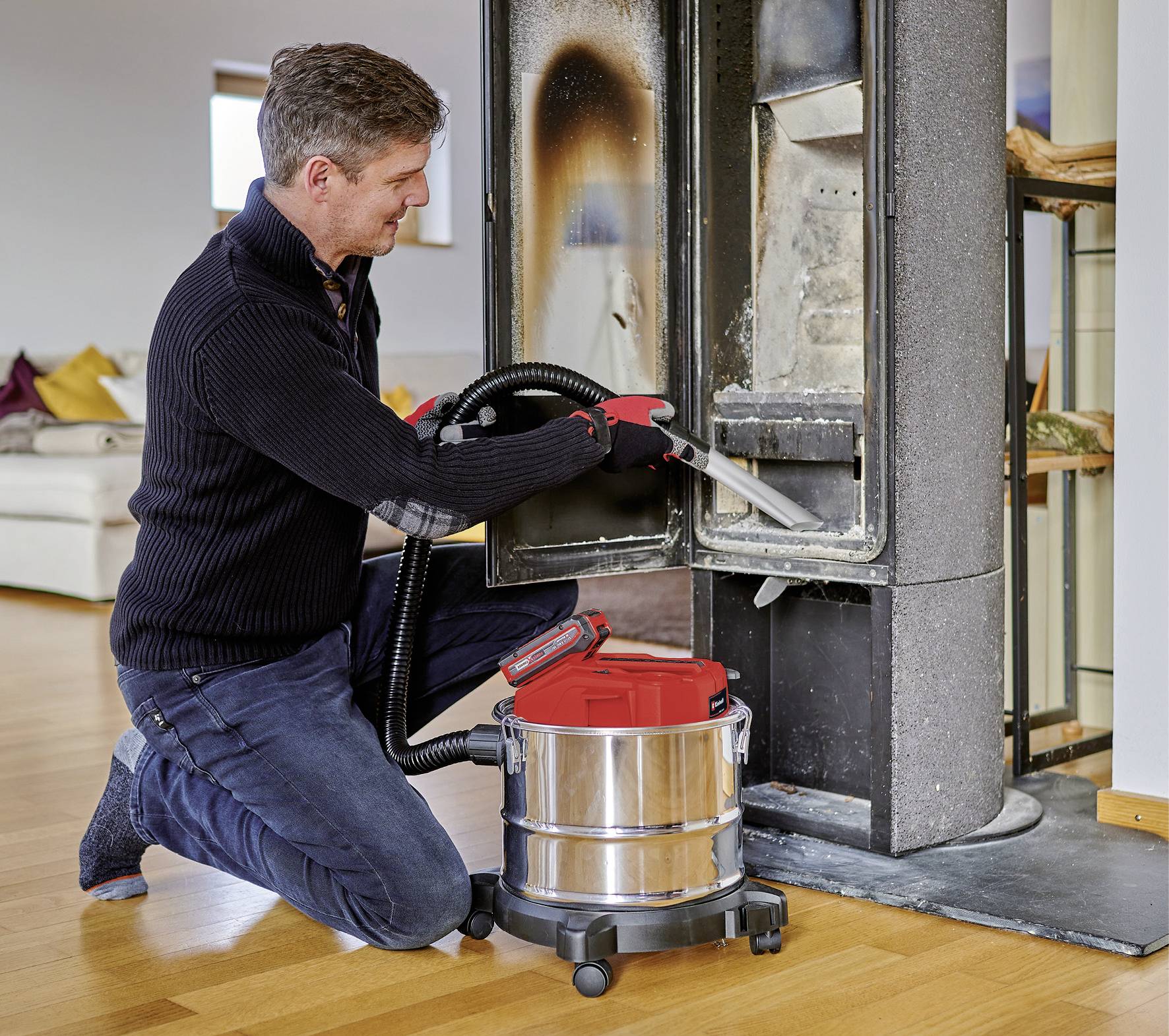 En man rengör en spis med en asksugare. Han bär svarta kläder och handskar, vardagsrummet syns i bakgrunden.