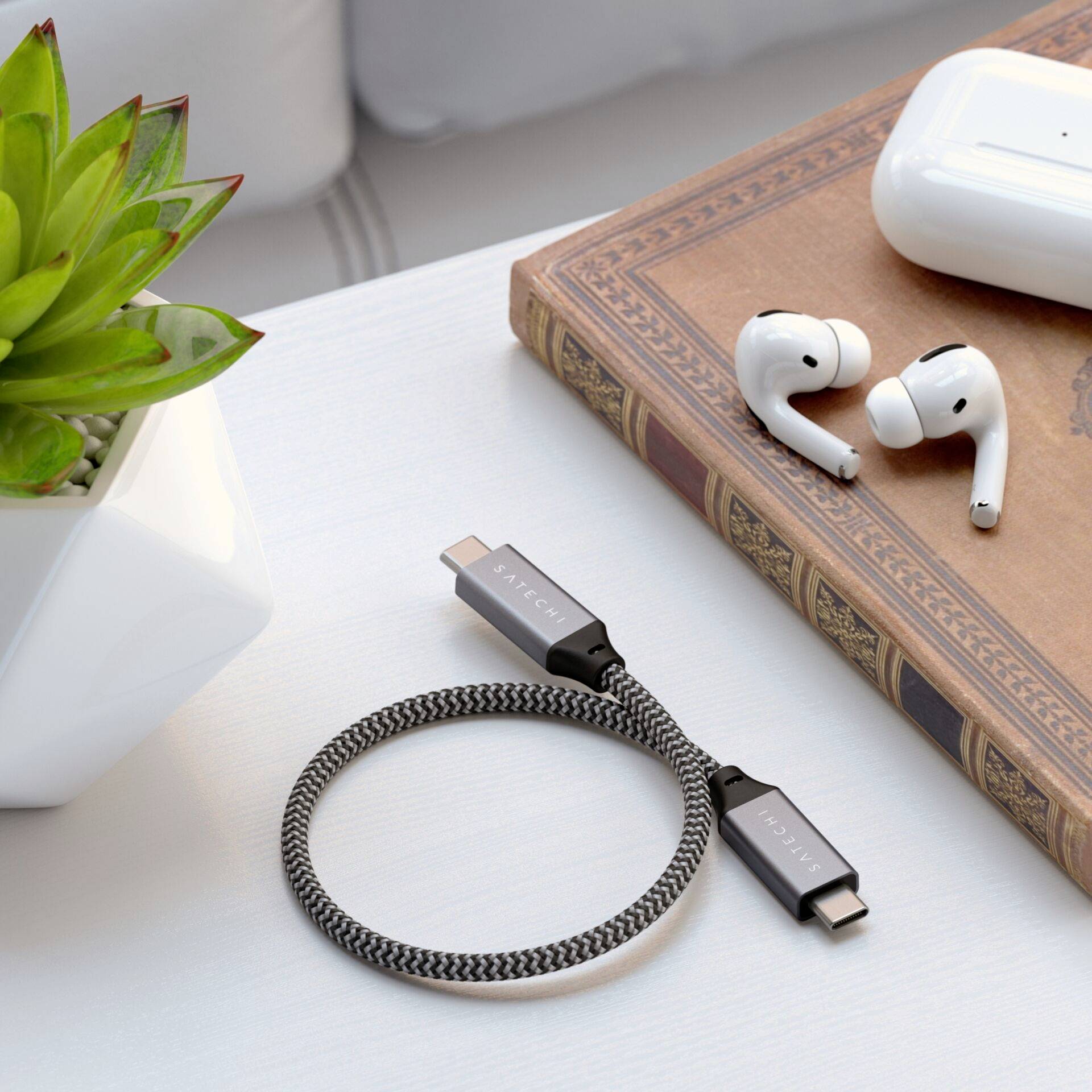 Ett laddningskabel, trådlösa hörlurar och en kruka på ett bord symboliserar teknik och natur i harmoni.