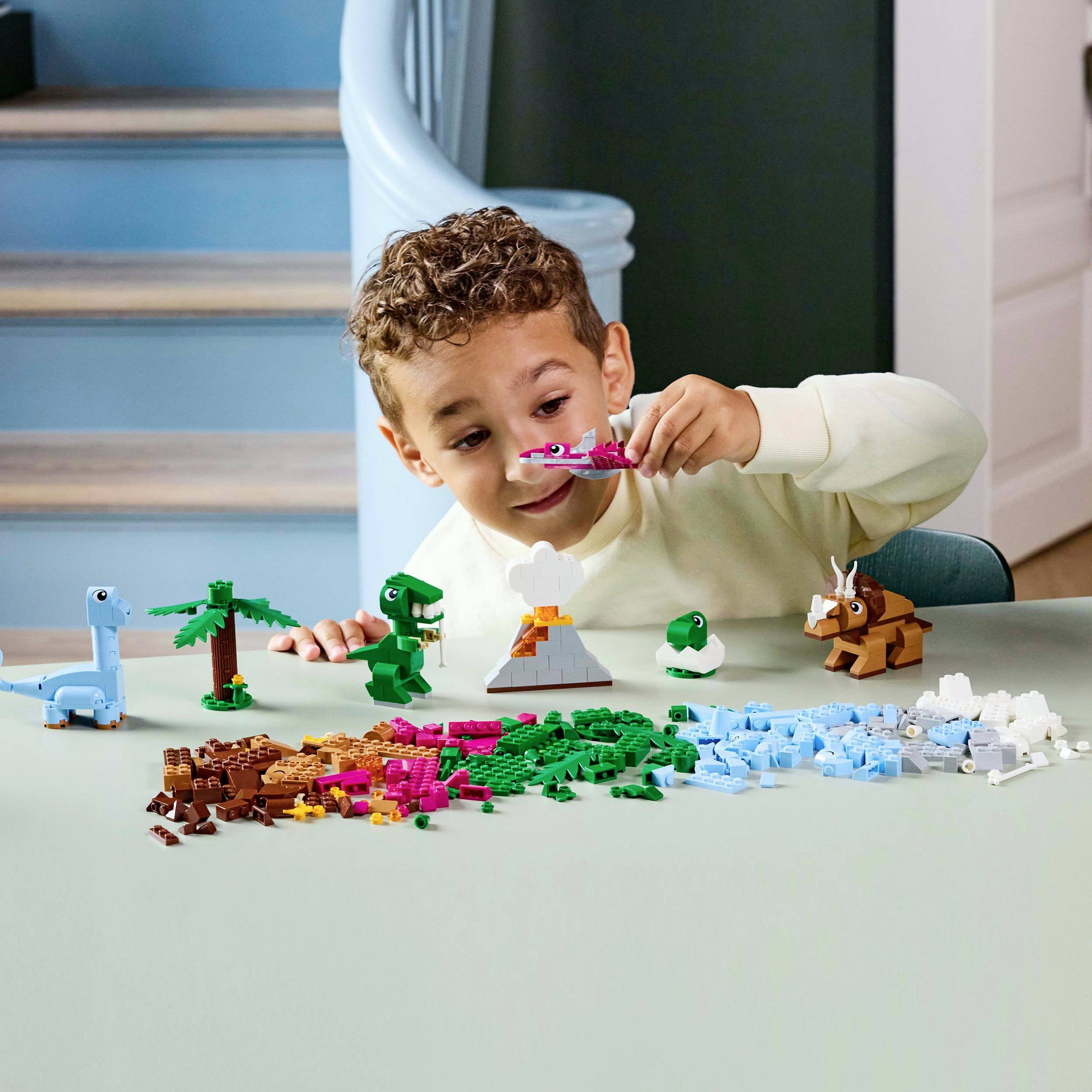 Ett barn leker med färgglada LEGO-dinosaurier på ett bord. I bakgrunden syns olika LEGO-djur och träd.