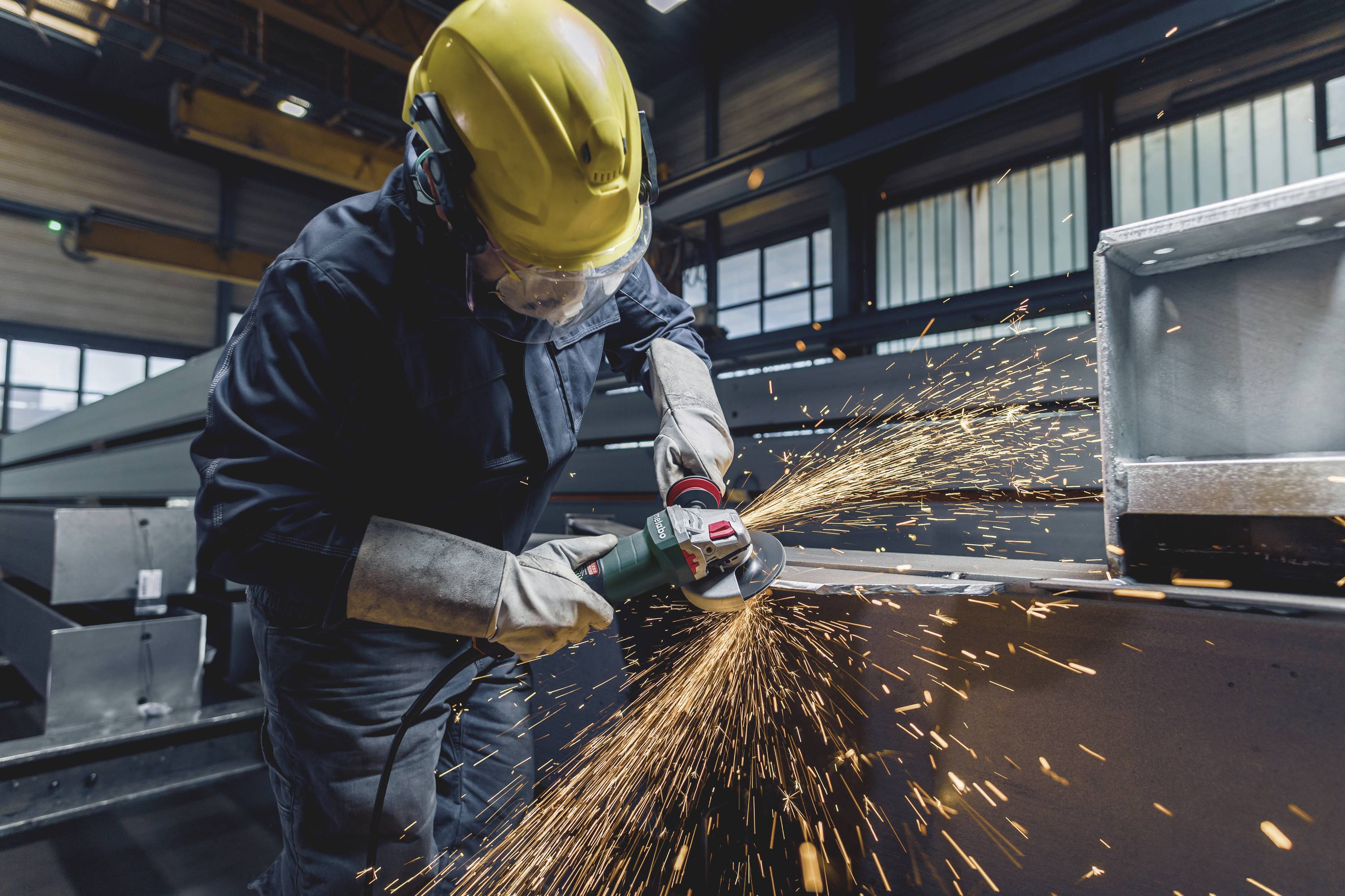 En arbetare bär skyddsutrustning och använder en vinkelslip för att ta bort metallsplitter. Produktionshall syns i bakgrunden.