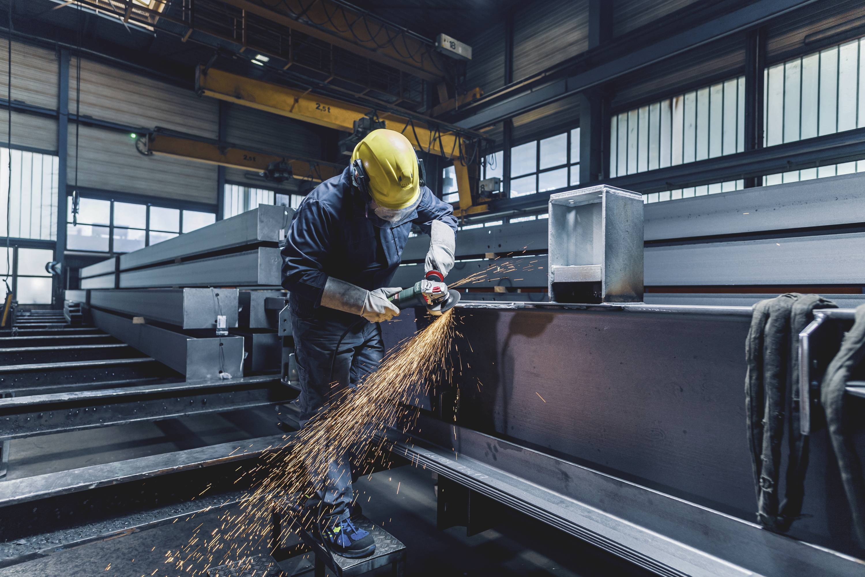 En arbetare med skyddshjälm bearbetar ett stort metallstycke med en vinkelslip i en fabrikshall, gnistor flyger.