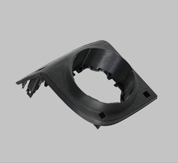 En svart plastdetalj som ser ut som en bilkomponent, möjligen en del av en fordonsklädsel, framför en grå bakgrund.