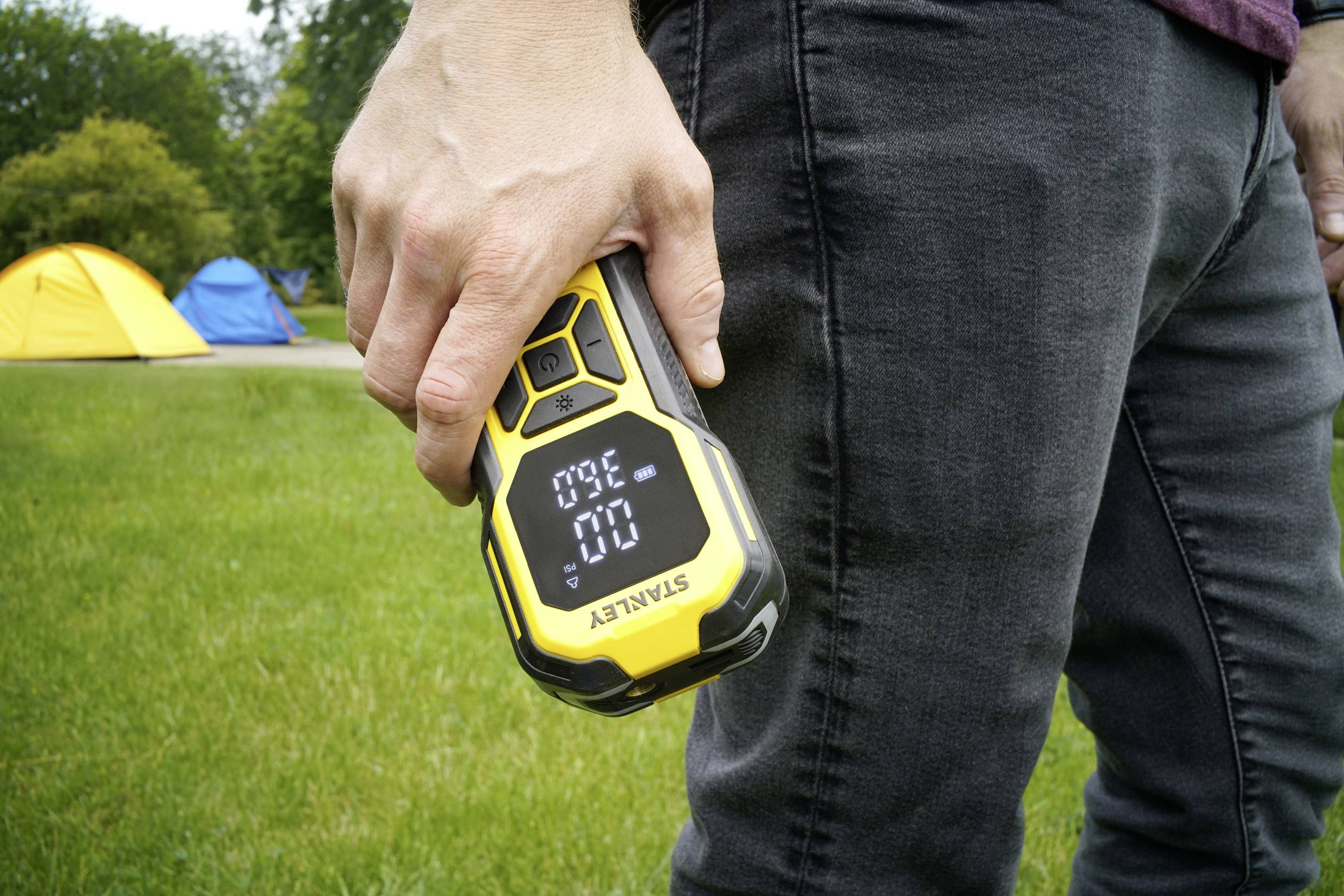 En person håller en gul enhet med en digital display som visar '30°' på en gräsmatta. I bakgrunden står två tält.