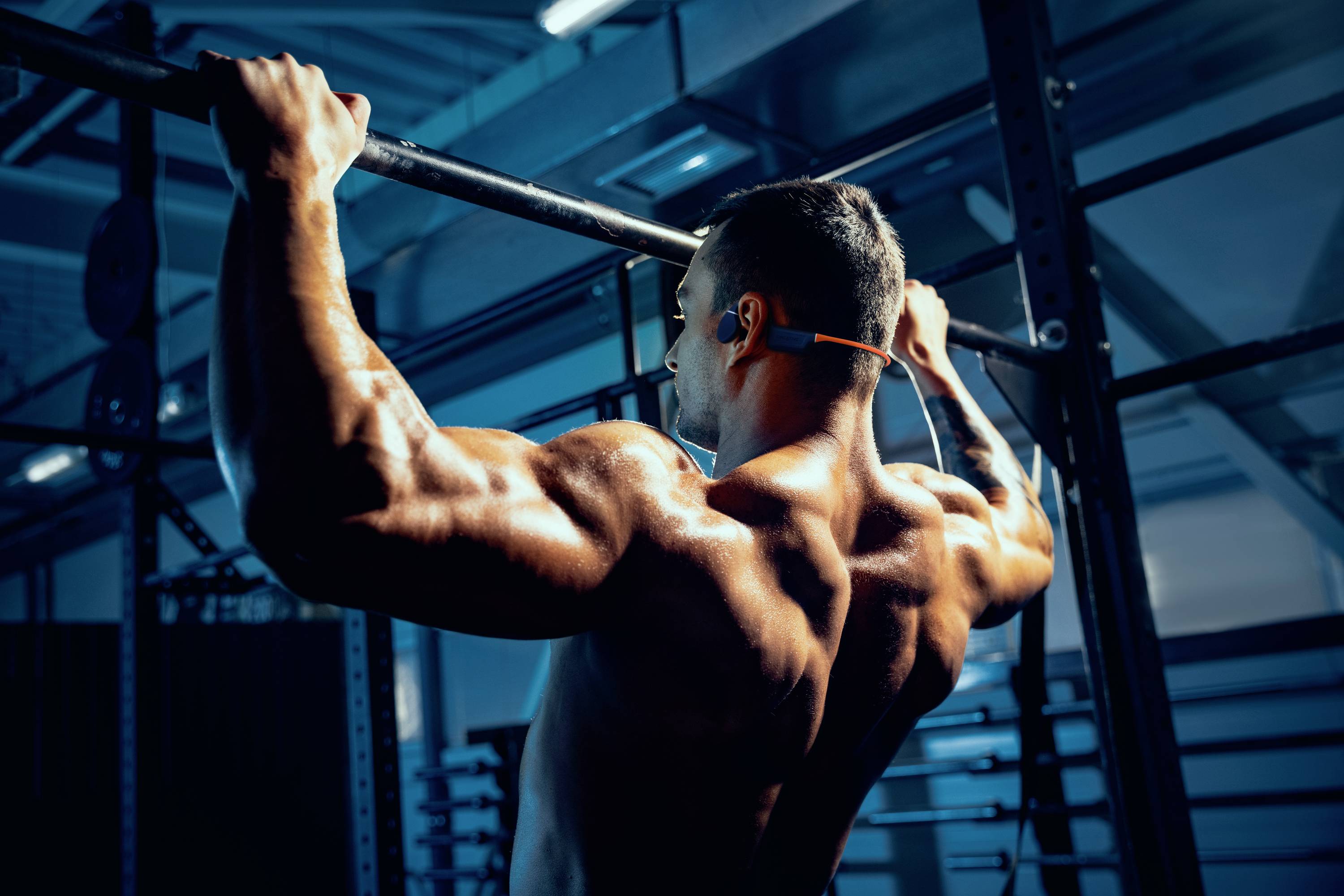 En muskulös man gör pullups i ett gym, belyst av blått ljus. Han bär trådlösa hörlurar och en tank top.