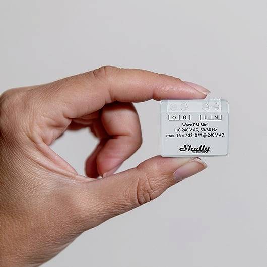 En hand håller en liten, vit enhet märkt 'Shelly'. Den visar symboler för elektriska anslutningar och spänningsspecifikationer.