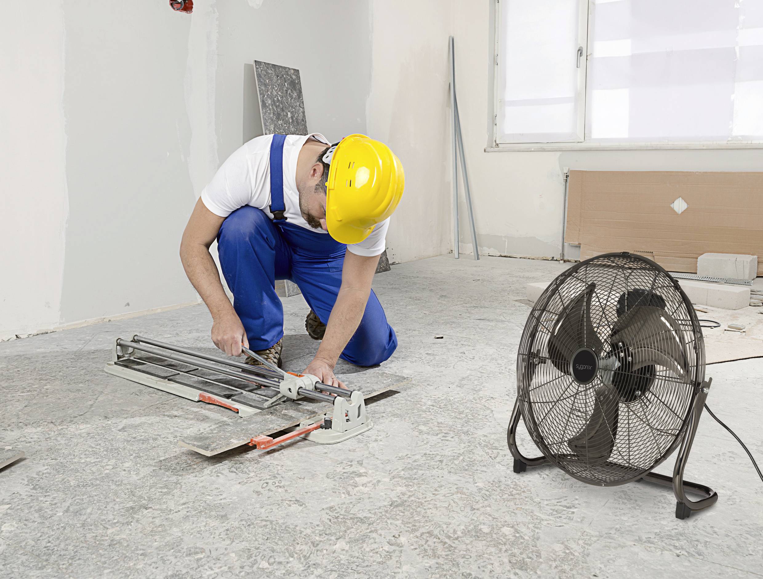 En arbetare med gul skyddshjälm skär kakel på ett golv i ett renoverat rum. En fläkt står bredvid.