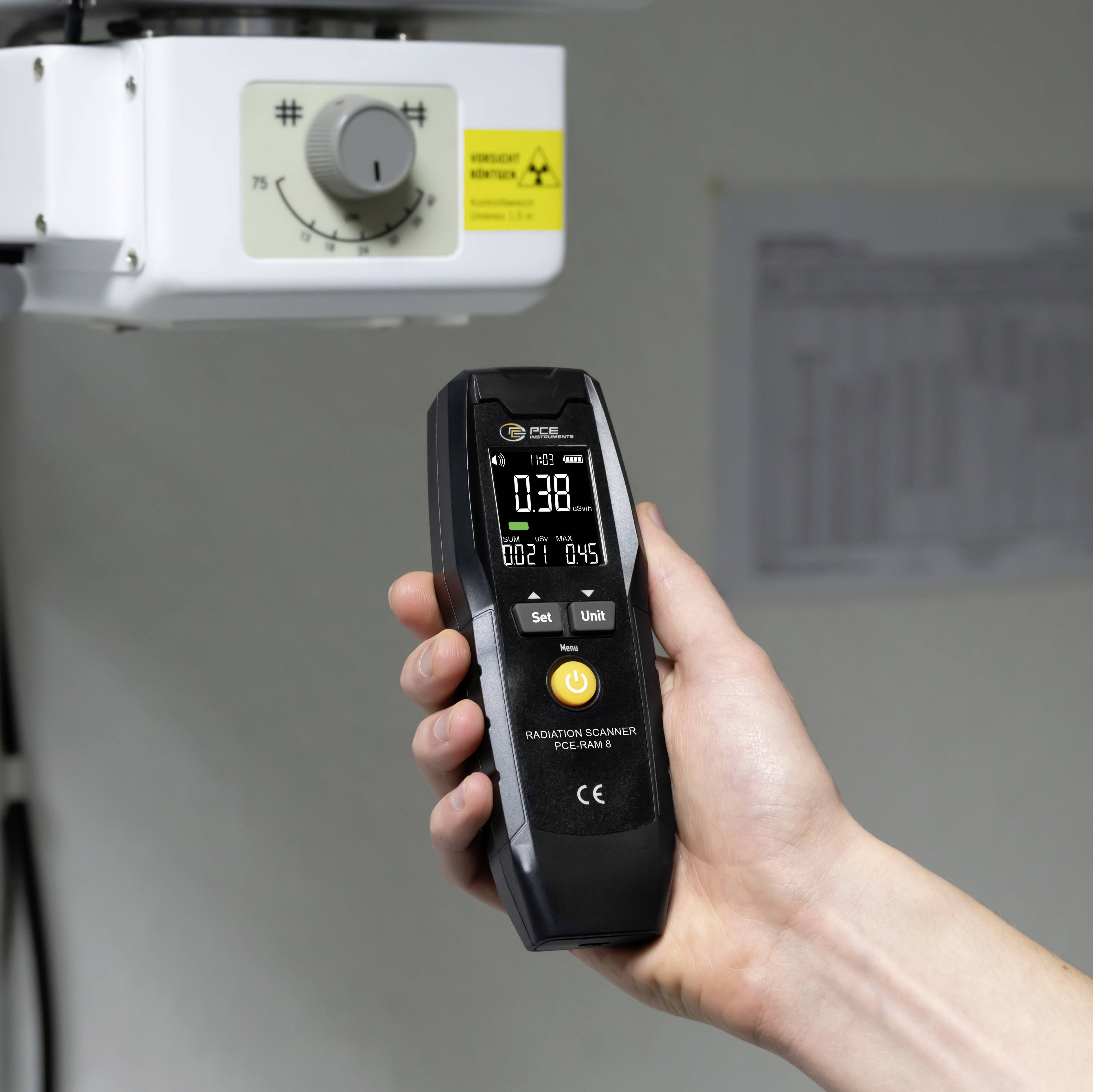 En hand håller en Geigermätare som under en röntgenapparatur visar en strålningsmätning på 0,38 µSv/h.