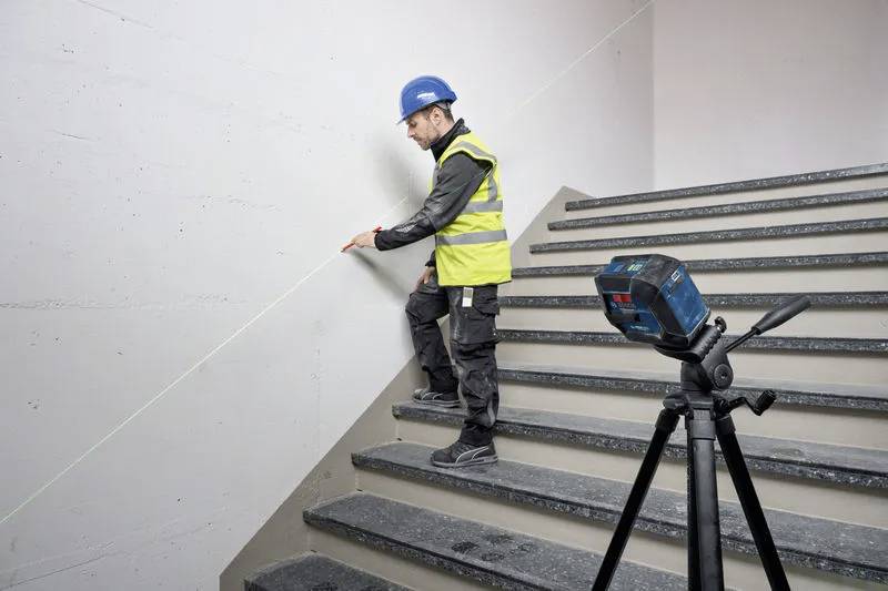En byggnadsarbetare med hjälm och varselväst använder ett lasernivåinstrument för att mäta vinkeln på en trappa. Lasern projicerar en grön linje på väggen.