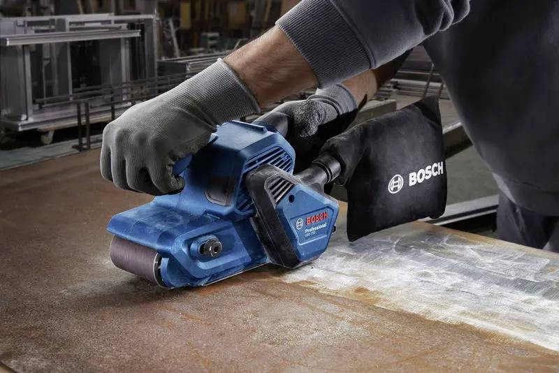 En person som använder en blå Bosch slipmaskin på en rostig metallyta, iklädd skyddshandskar.