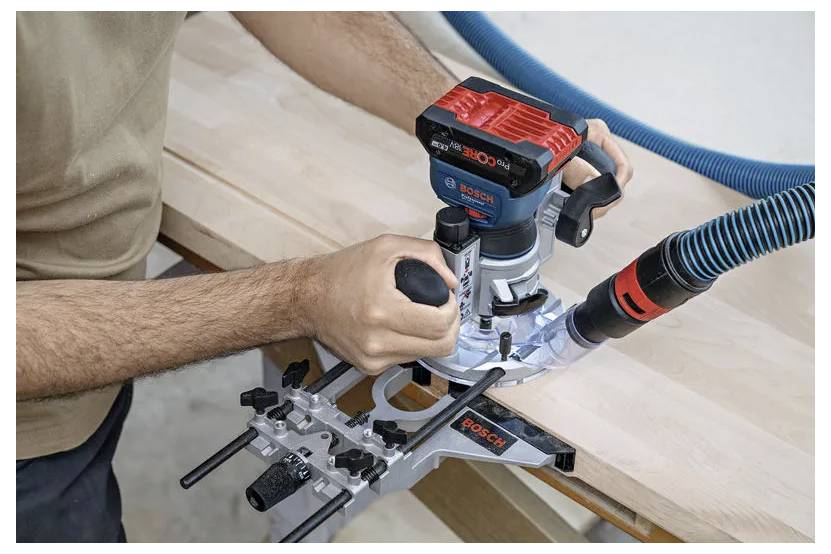 En person använder en Bosch-router på en träyta, med en blå vakuumslang ansluten, och styr den längs kanten för precision vid formgivning.