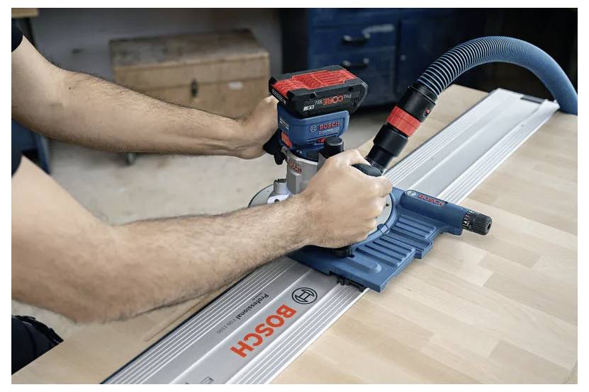 En person använder en Bosch-router på en träyta, guidad av en metallskena. Arbetsytan har verktyg och lådor i bakgrunden.