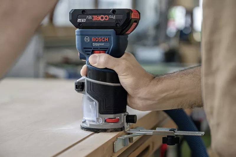En person använder en Bosch elektrisk fräs längs en träkant för att utföra precisionsarbete. I bakgrunden en oskarp verkstadsmiljö.