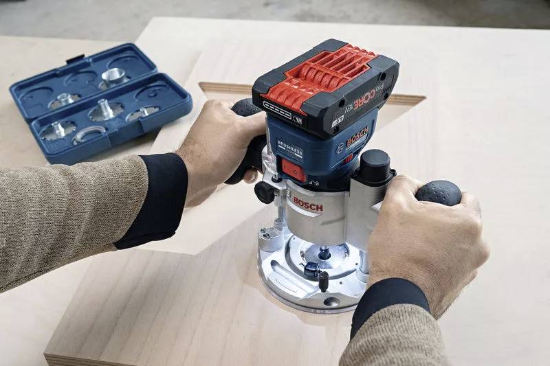 En person använder en Bosch-fräs på ett träbord. I bakgrunden syns en öppen väska med fräsinsatser.