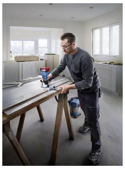 En man använder en elborr för att montera eller reparera ett träarbordsbord i ett ljust, obehandlat rum.
