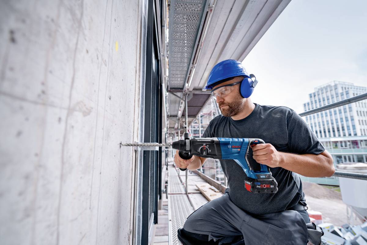 En byggnadsarbetare med skyddshjälm och hörselskydd borrar hål i en betongvägg på en byggarbetsplats med en elektrisk borr.
