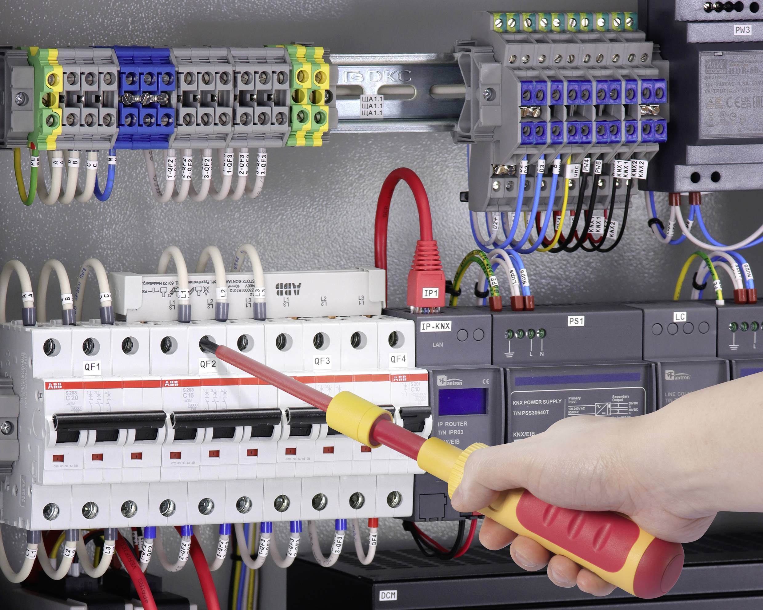 En person arbetar med en skruvmejsel på en kontrollpanel med många elektriska kretsar och färggranna kablar.