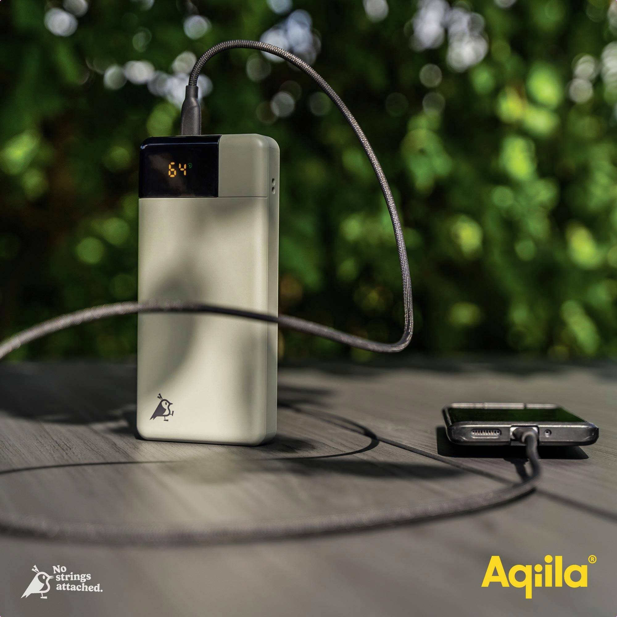 En powerbank laddar en smartphone via en kabel. På powerbanken visar en display '64', sannolikt batterinivån. I bakgrunden ett oskarp grönt fält.