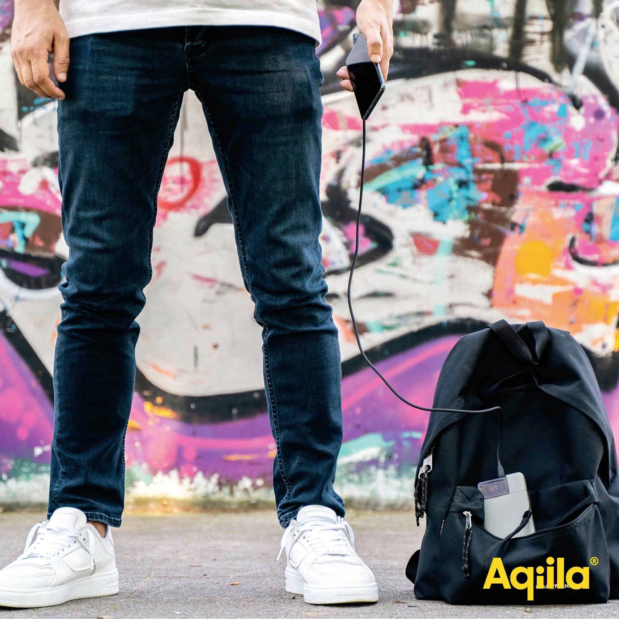 En person håller en mobiltelefon som är ansluten till en powerbank i ryggsäcken framför en färgglad graffitivägg.