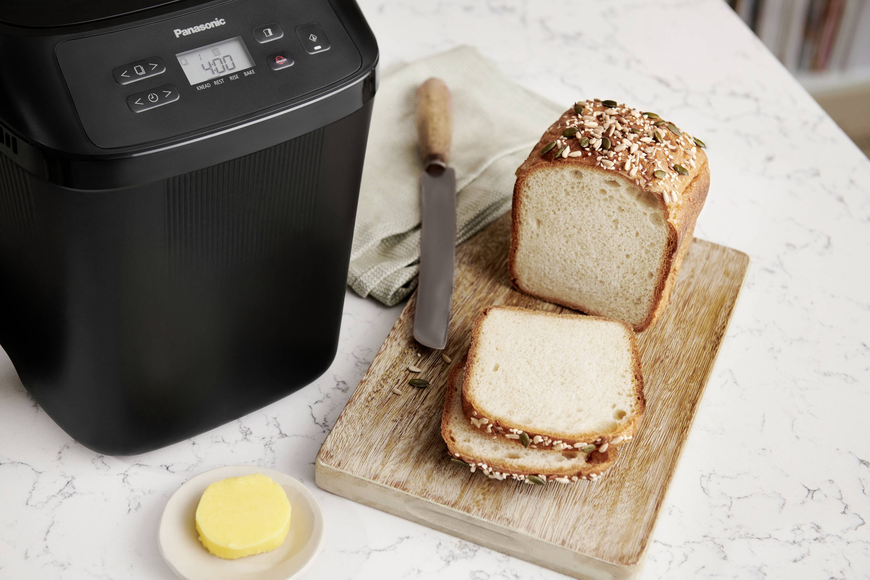 En brödbakningsmmaskin på ett köksbord, bredvid ett skärbräde med skivat bröd. En kniv och smör syns också.