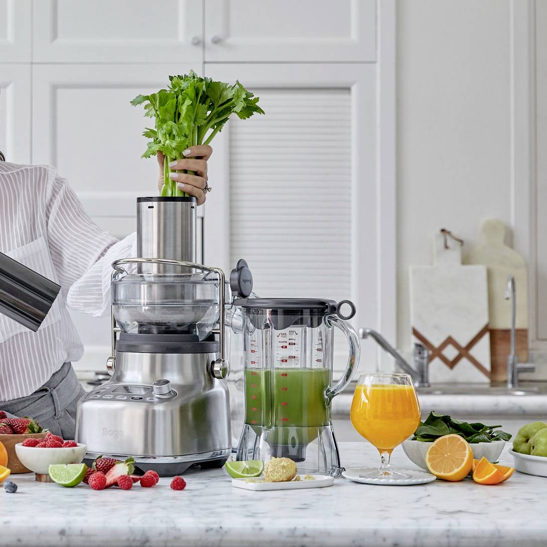 En person blandar grönsaker och frukt i en juicemaskin i ett kök. På bordet ligger bär, limefrukter, apelsiner och ett glas juice.