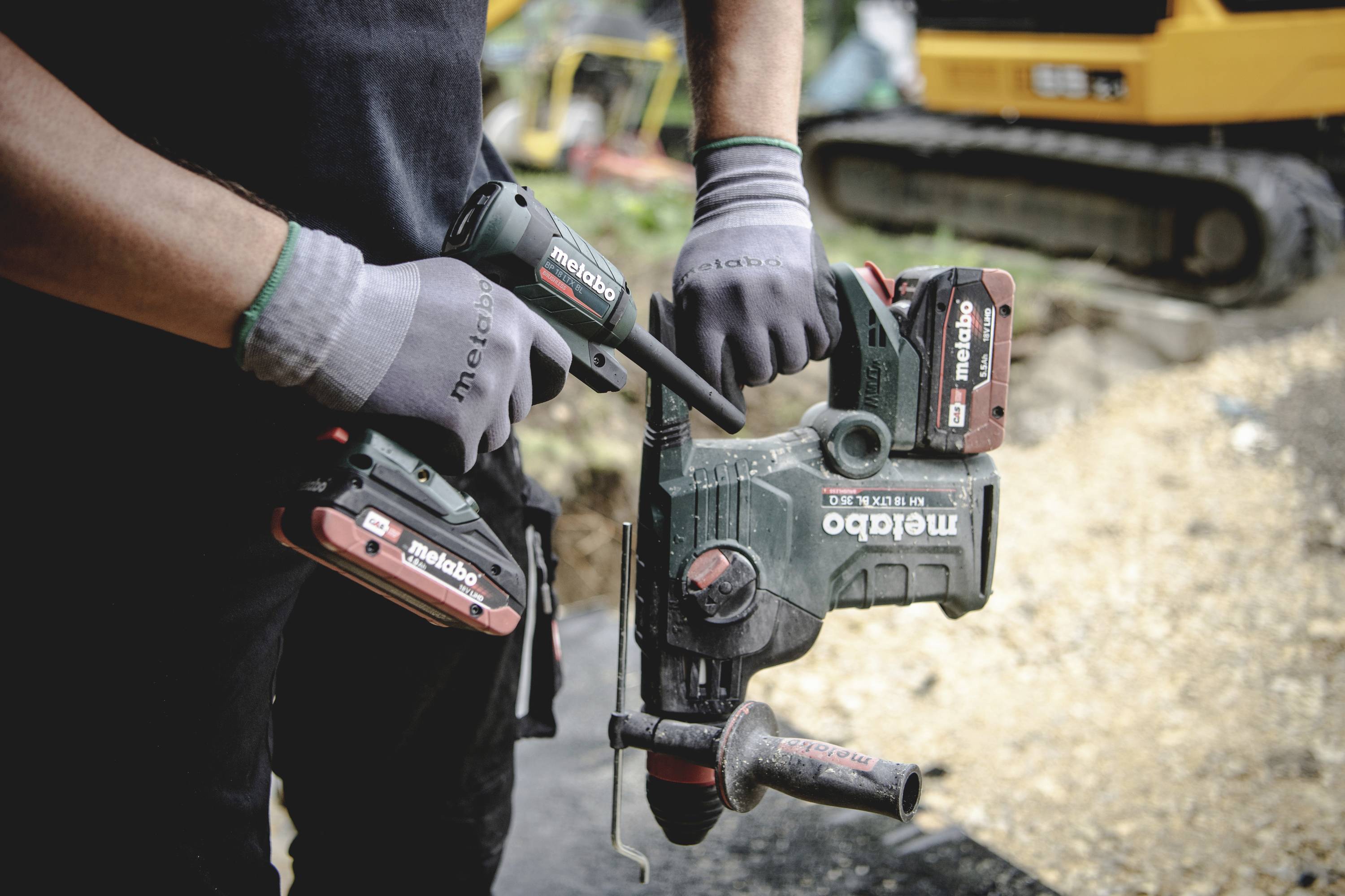 En person håller en Metabo-borrhammare med batterier, bär handskar, byggarbetsplats i bakgrunden, grävmaskin synlig.