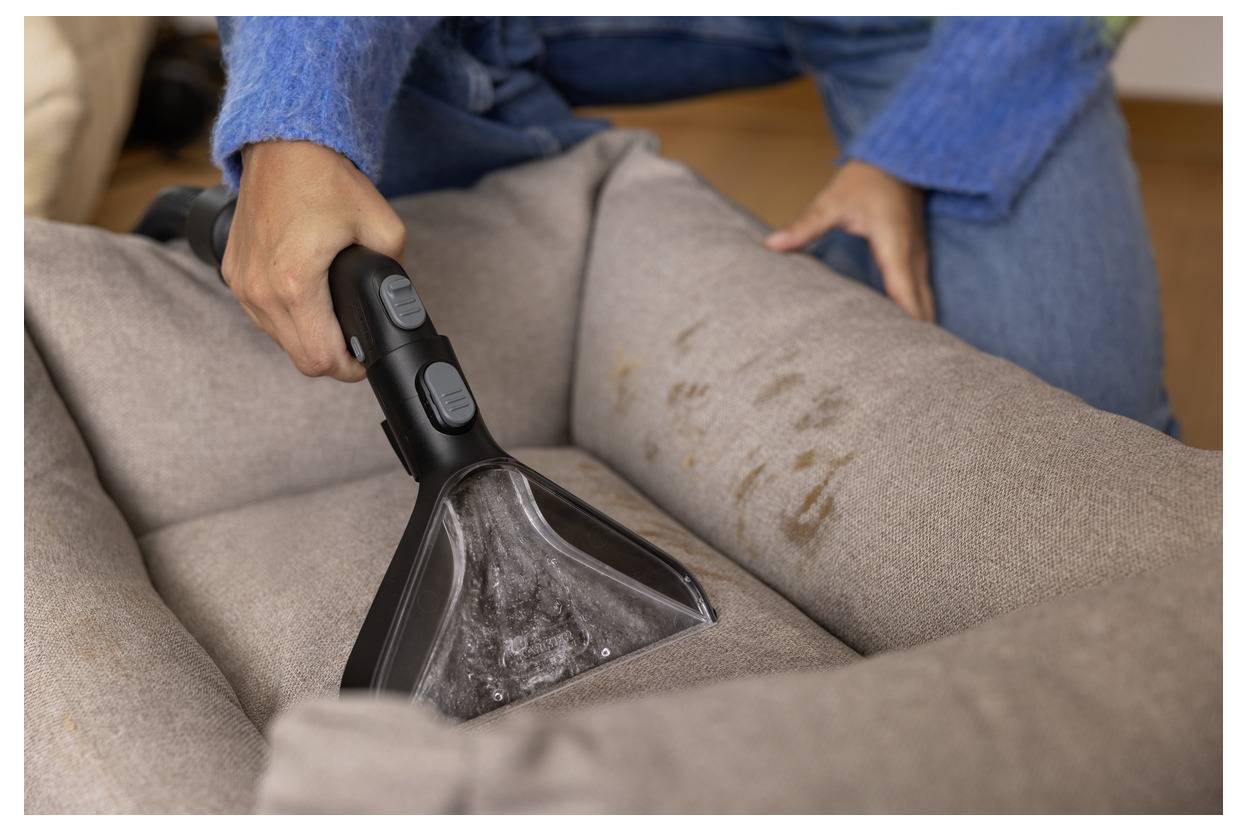 En person använder en handhållen dammsugare med borstatillsats för att rengöra en djursäng med synliga fläckar, klädd i en blå tröja och jeans.